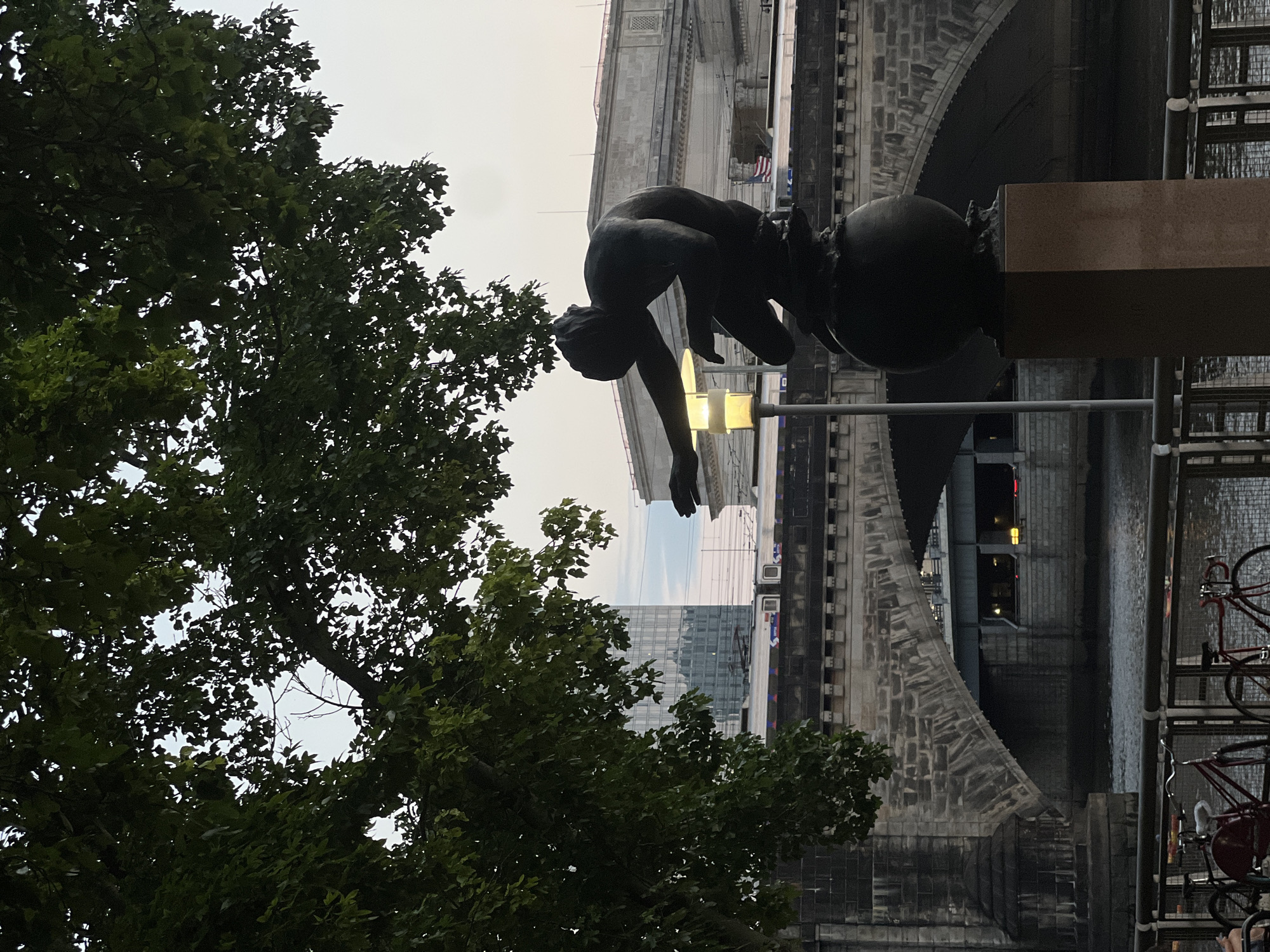 The image shows a bronze sculpture of a male figure kneeling on a globe, with its right arm extended forward. The sculpture is silhouetted against a cloudy sky and is positioned in front of a stone bridge that spans a body of water. In the background, a train is visible on tracks that run across the bridge, and beyond that, a modern glass-facade building stands tall. The scene is framed by the dark green leaves of a large tree in the foreground. This location is the Benjamin Franklin Parkway in Philadelphia, Pennsylvania, USA, with the sculpture likely being part of the city's many public art installations. The atmosphere suggests it is either early morning or late evening, given the lighting conditions. The scene is devoid of any prominent human activity beyond the potential implied presence of the train passengers.