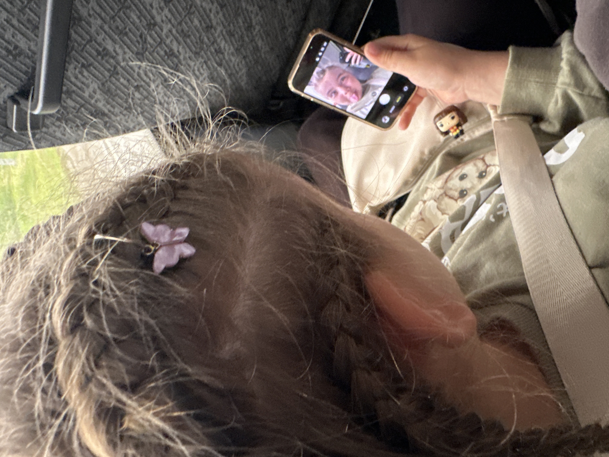 The image displays a close-up, over-the-shoulder view of a child seated within a vehicle, observed from the child's right side. The provided image does not contain visual information to confirm the specified location in the city of Кварса, Russia.
The child has light brown hair styled in braids, with numerous fine, loose strands. A small, light purple butterfly-shaped hair clip is affixed to the hair on the right side of the child's head. The child's right ear is visible. The child is wearing a light green long-sleeved top featuring a subtle pattern, possibly of animal faces or similar motifs. A light-colored seatbelt diagonally crosses the child's torso.
The child's right hand holds a smartphone with a metallic gold or bronze frame. The phone's screen displays a front-facing camera view, showing the reflection of a person, possibly an adult, with light-colored hair, making a playful facial expression with their tongue extended. On the child's lap or clothing, below the phone, a small Funko Pop figurine with brown hair and a striped top is visible.
The background indicates an interior setting, likely a vehicle such as a car or bus. To the left, a window shows blurred green foliage and bright daylight. To the right, a dark, textured interior panel of the vehicle with a visible dark handle is present. The seating material behind the child appears to be a dark, textured fabric.