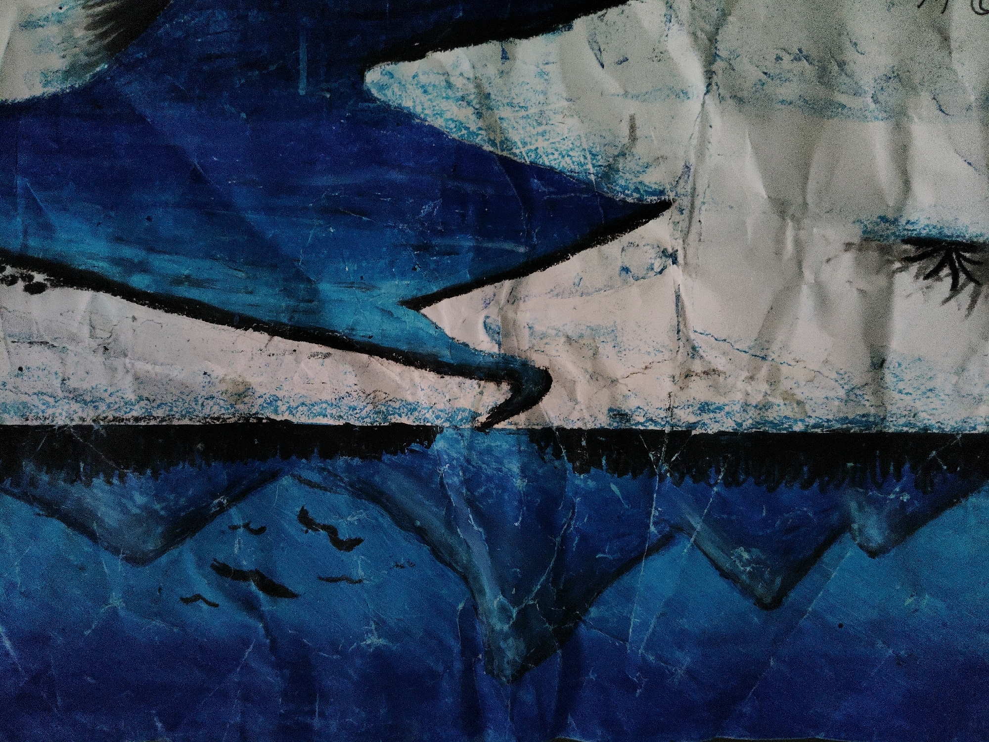 The image displays a close-up, abstract painting with a prominent use of blue and white, likely depicting a landscape with water and mountains. The texture of the paper is visible, with wrinkles and folds affecting the painted surface. In the lower portion of the painting, dark, triangular shapes suggest mountains reflected in a body of deep blue water, with a few small black shapes possibly representing birds in flight. The upper portion features broader strokes of blue and white, with black lines forming sharp angles, perhaps representing landforms or abstract shapes. There is a suggestion of a dark, jagged line separating the upper and lower sections, possibly depicting a horizon. The painting has a raw, textured quality, as if created with thick paint or pastels on crinkled paper. There are no discernible elements that would indicate a specific location in Duminichowki, India, nor are there any people, specific objects, or actions depicted.