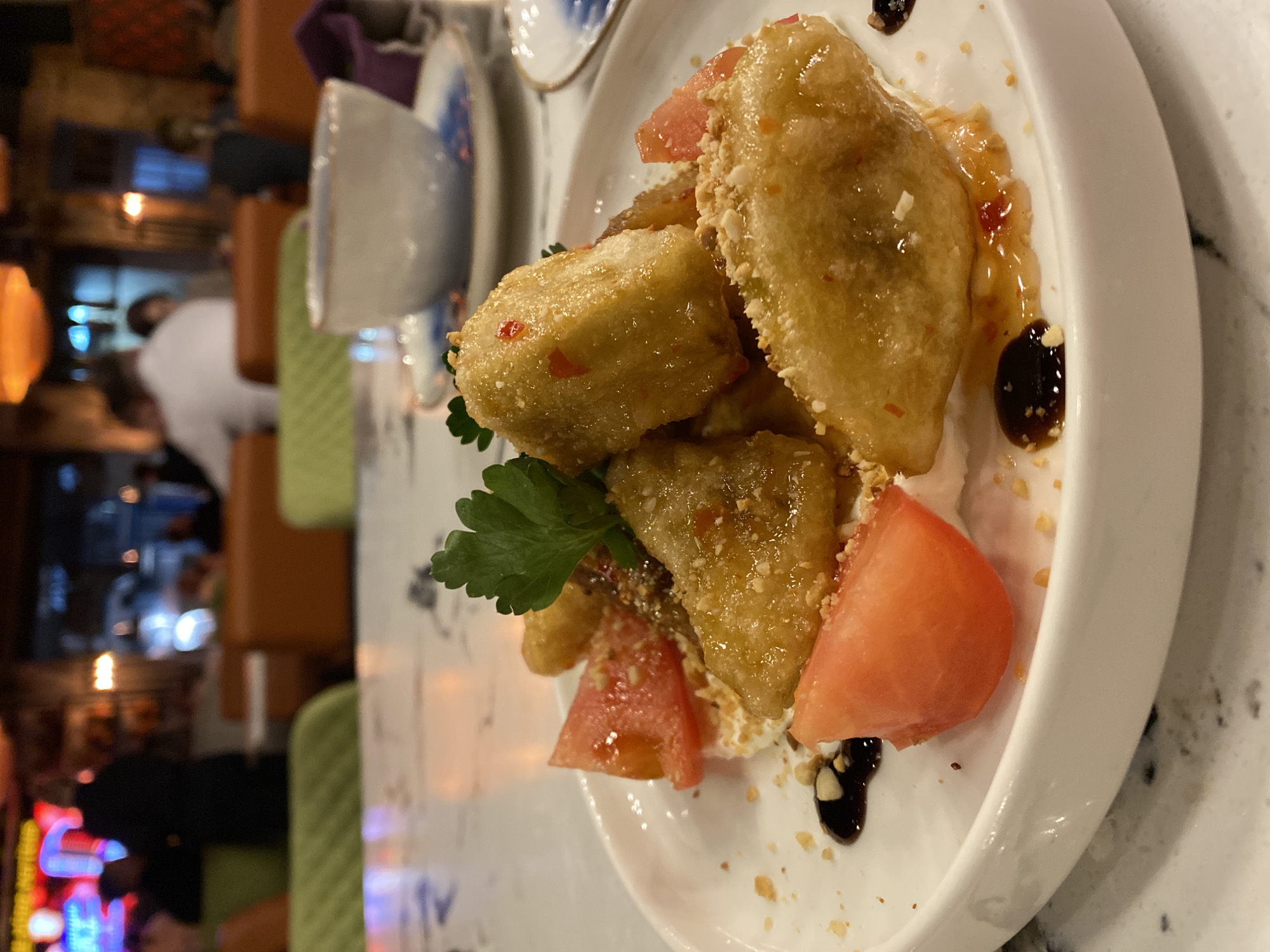 The image shows a plate of food, likely in a restaurant. The main dish appears to be fried or battered tofu, served with sliced tomatoes, parsley, and a sweet sauce or glaze. The restaurant environment is visible in the background, with tables, chairs, and other patrons present. The setting suggests a somewhat upscale establishment with warm lighting and decor. I lack access to data that would allow me to identify the location as being in Yekaterinburg, Russia.