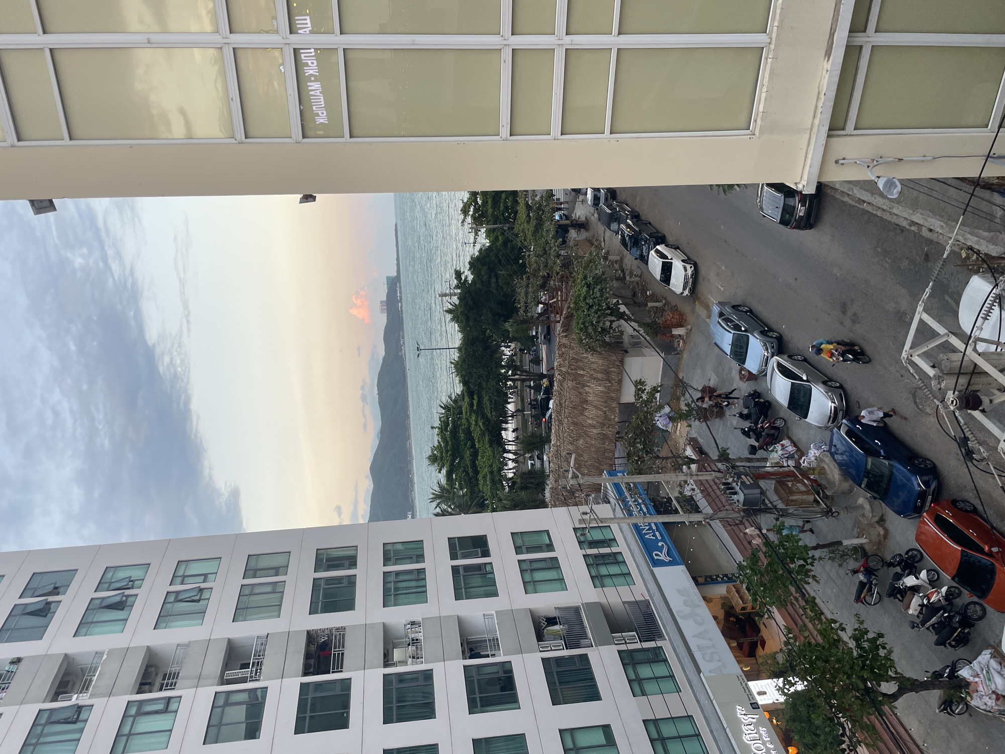 The image shows an elevated view of a street in Bac Nha Trang Ward, Vietnam, overlooking the ocean. The scene is captured between two buildings, one beige with multiple window panes, and a taller, white residential building with balconies. Below, the street is lined with parked cars and motorbikes. Several people are present, some appearing to be standing near the vehicles. There's a thatched roof structure near the road, possibly a small kiosk or shelter. In the background, the ocean meets the shore, with landforms visible in the distance under a cloudy sky. A sign for a hotel or business is visible along the side of the white building.