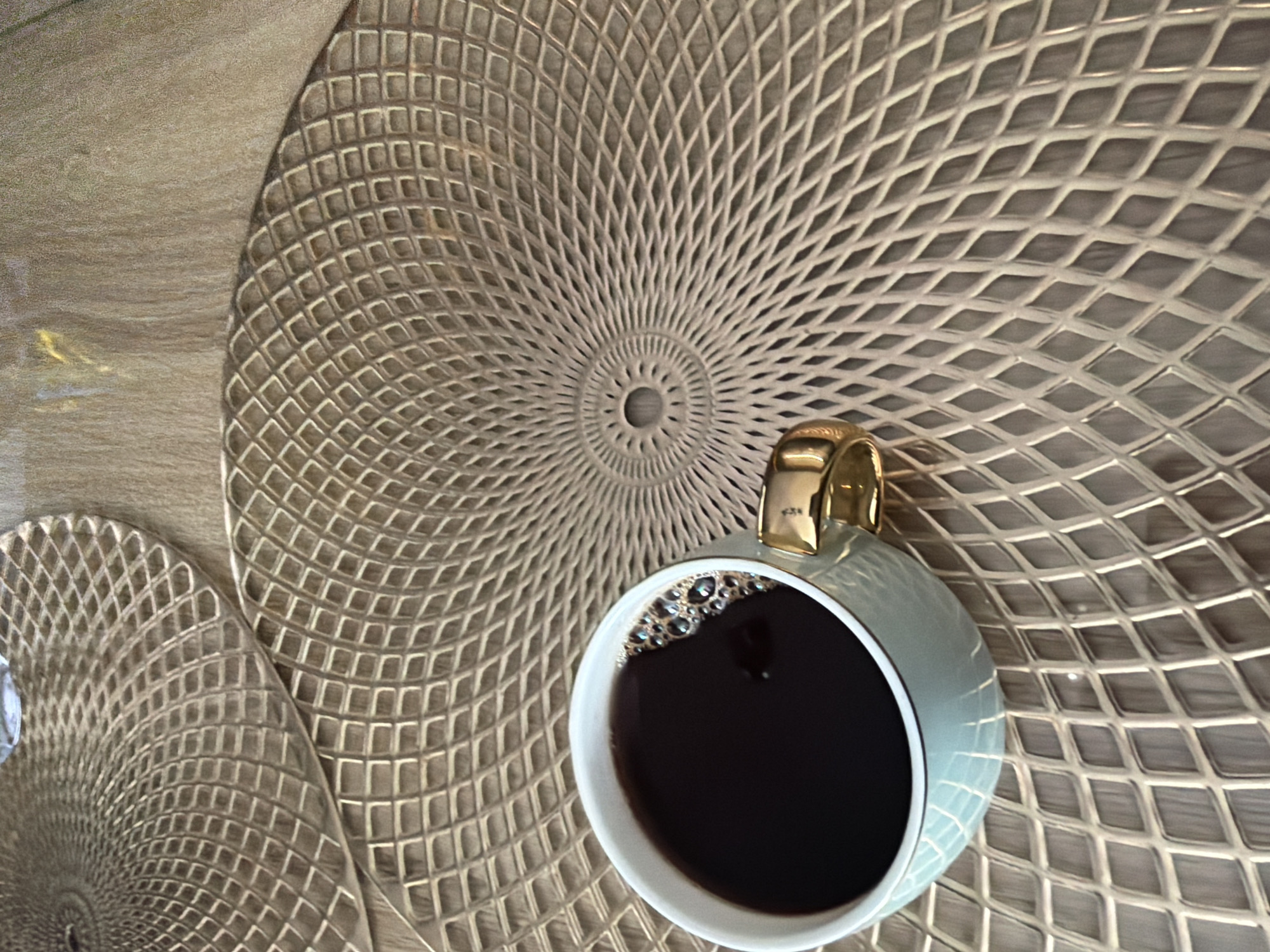 An overhead shot shows a light blue coffee cup filled with dark coffee and a golden handle, resting on a gold, diamond-patterned placemat. A second, similar placemat is visible in the upper left corner of the image. The placemats are on a wooden surface. The scene depicts a still life of coffee and placemats on a table.