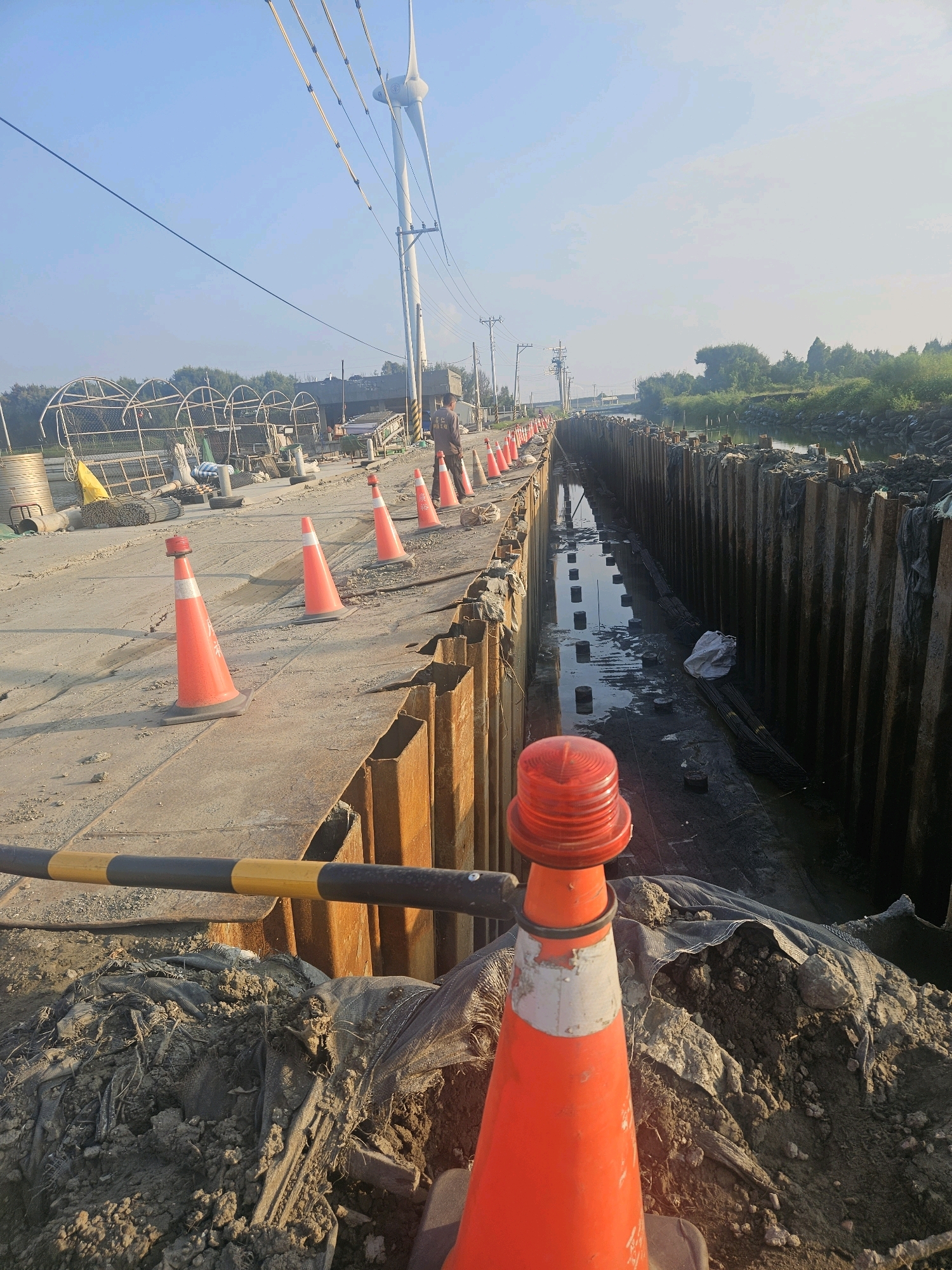 In Taixi Township, Taiwan, a construction site is depicted. A trench, possibly for utility work, has been excavated and is lined with sheet pilings. The trench is partially filled with water and mud, with rebar and wooden stakes visible on the trench floor. Construction cones, including one with a flashing orange light, are placed along the edge of the trench, marking a boundary. A black and yellow striped barrier is also present. In the background, a wind turbine stands tall under a clear blue sky. Several industrial structures and vegetation line the perimeter of the site. A person is visible in the distance, observing the construction. The overall scene suggests ongoing infrastructure development.