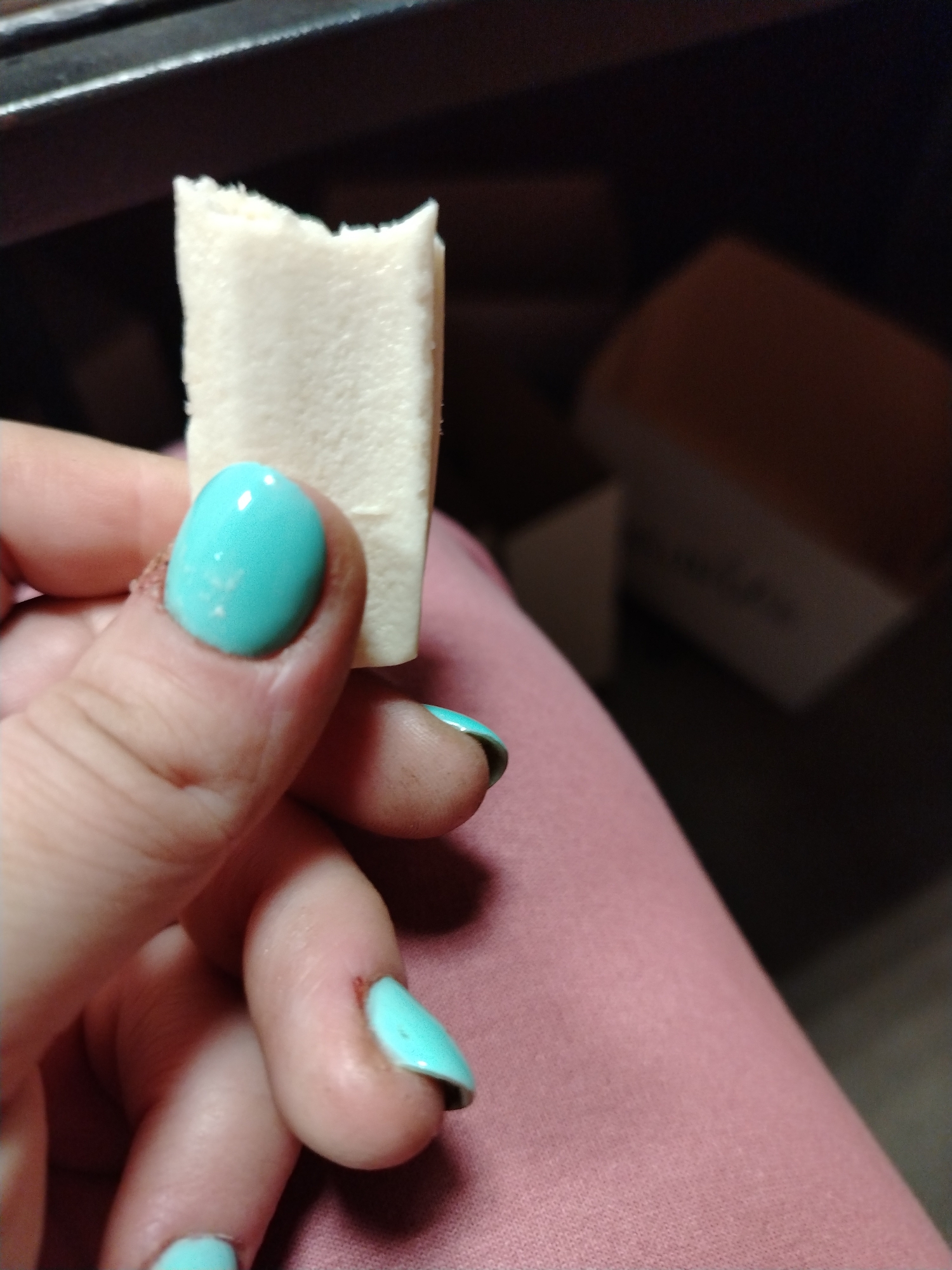 The image captures a close-up of a person's left hand holding a rectangular, off-white food item, which appears to have a bite taken from one end. The food item has a soft, somewhat porous texture along its bitten edge. The hand features bright turquoise (teal) nail polish on the fingernails. Some of the nail polish, particularly on the thumb and ring finger, shows signs of chipping or wear. The skin around the nails also appears to have minor imperfections.
The background is dim and out of focus. In the upper left, a dark, indistinct surface, possibly a piece of furniture or shelving, is visible. To the right, a soft, light pink fabric, likely clothing or a blanket, fills a portion of the frame. Further in the blurred background, there is a light-colored box with dark, indistinct text printed on it, which is too blurry to be precisely identified but might suggest words like "Chocolate." The setting appears to be an indoor, casual environment. The image is taken in Шебунино, Russia.
