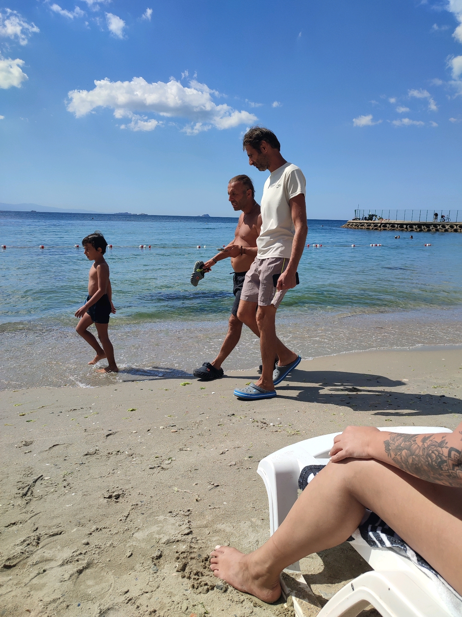 On a sunny day at Moda Beach in Kadıköy, Istanbul, Turkey, three people are present. In the foreground, a person is reclining on a white beach chair, their tattooed arm and legs visible. Further back, walking along the shoreline, are two adult males and a young boy. The boy, wearing dark blue swimming trunks, is wading in the shallow water, while the two men walk beside him on the wet sand. One man, shirtless, carries a pair of grey sandals in his hand, and the other, wearing a light-colored t-shirt and shorts, is holding a dark object, possibly a phone. The sea is calm and blue, with buoys marking a swimming area. In the background, a pier extends into the water, with structures and people visible on it. The sky is bright blue with scattered white clouds.