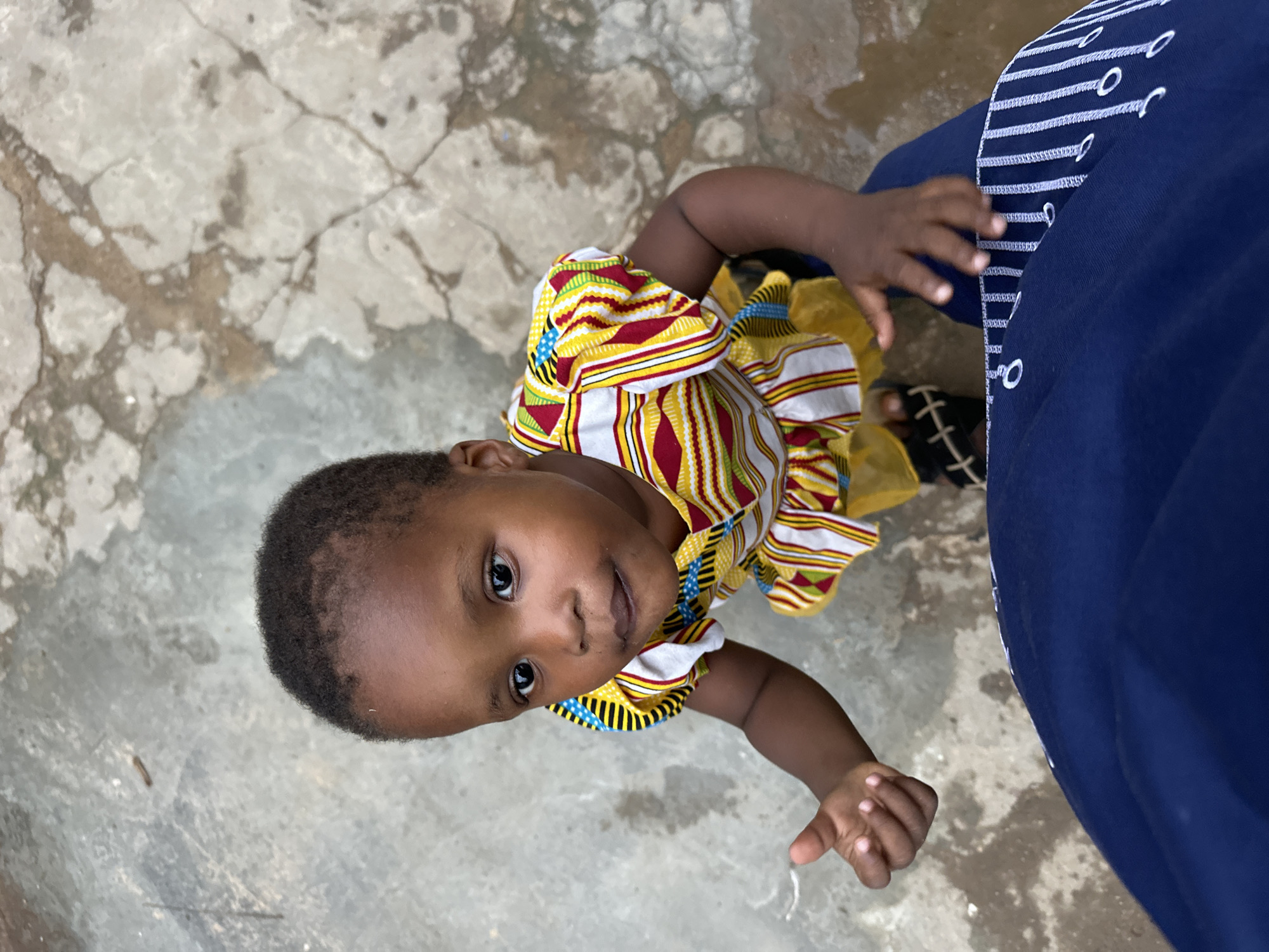 In Narayi, Nigeria, a young child is captured from a high-angle perspective, looking upwards with a curious expression. The child is dressed in a vibrant, multi-colored striped dress with yellow and red accents, featuring ruffles at the hem. The child's skin is dark, and their hair is short and dark.
The setting appears to be outdoors, with a textured, concrete ground that shows signs of wear and discoloration. To the right of the child, a person is partially visible, wearing a navy blue garment with white embroidered lines and circles along the edge. The child's small hand is reaching out towards this person. A sandal with a black strap and a white crisscross pattern is visible on the ground near the person's feet.
The lighting suggests it is daytime, and the overall mood of the image is one of innocent curiosity and interaction. The composition focuses on the child's face and their engagement with the unseen person. No visible text is present in the image.