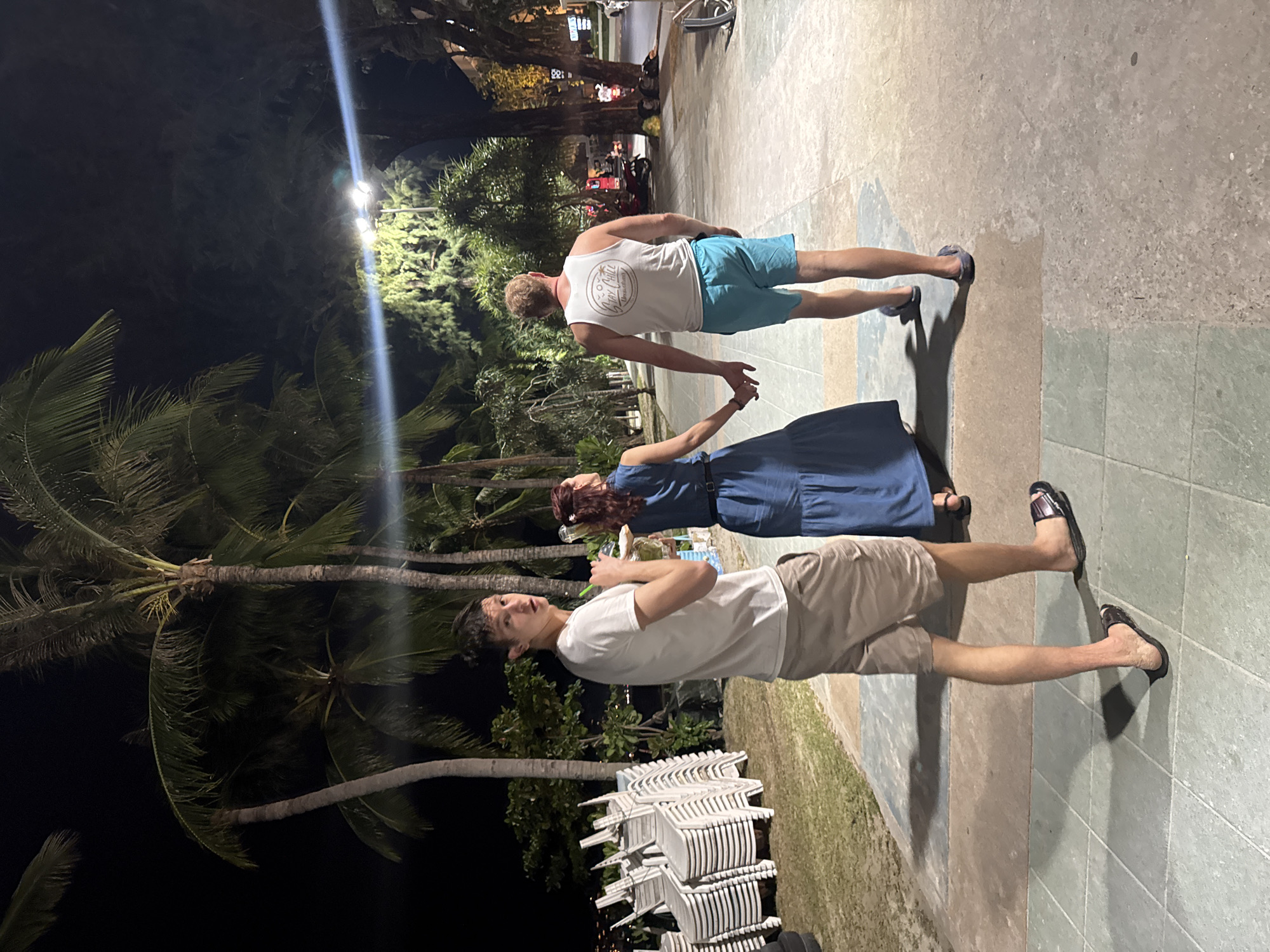 The image shows three people standing on a tiled walkway near a beach at night. The location appears to be Patong, Thailand, based on the presence of palm trees, stacked white plastic chairs typical of beach setups, and the overall ambiance.
From left to right: A man in a white t-shirt and khaki shorts is standing near a stack of chairs and appears to be holding a small glass or bottle. A woman in a long, dark blue dress is holding hands with another man, who is wearing a white tank top with text and light blue shorts. The background features palm trees and a dimly lit street with some traffic. The area is illuminated by streetlights and possibly lights from nearby establishments.