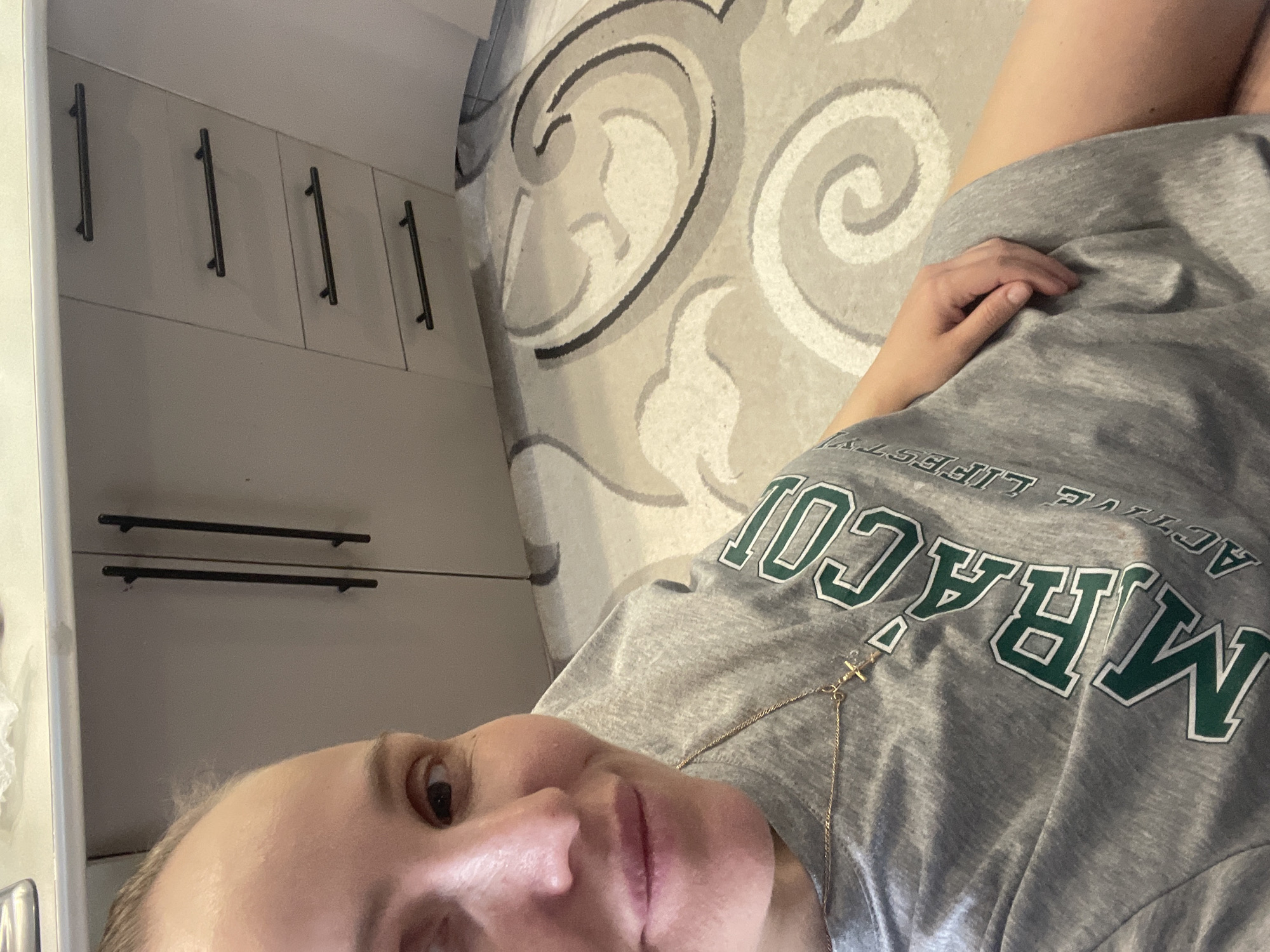 A selfie captures a person reclining indoors, looking directly at the camera with their eyes open. The image is rotated, showing the person on their side. They wear a casual grey t-shirt featuring green and white text that reads "MIRACULOUS ACTIVE LIFE" (visible upside down from the viewer's perspective). A delicate gold chain with a small cross pendant hangs around their neck. One hand is resting on their mid-section, and part of a bare leg is visible.
The background reveals a patterned rug with swirling designs in shades of grey, white, and black, suggesting it's either on the floor or a large mat. To the left, light-colored cabinets with modern, dark, horizontal bar handles are visible. These cabinets appear to be part of a kitchen or storage area. The lighting is consistent with an indoor setting, possibly during the daytime or under ambient artificial light. The overall mood is relaxed and personal. This scene is situated in Chaikovsky, Russia.
