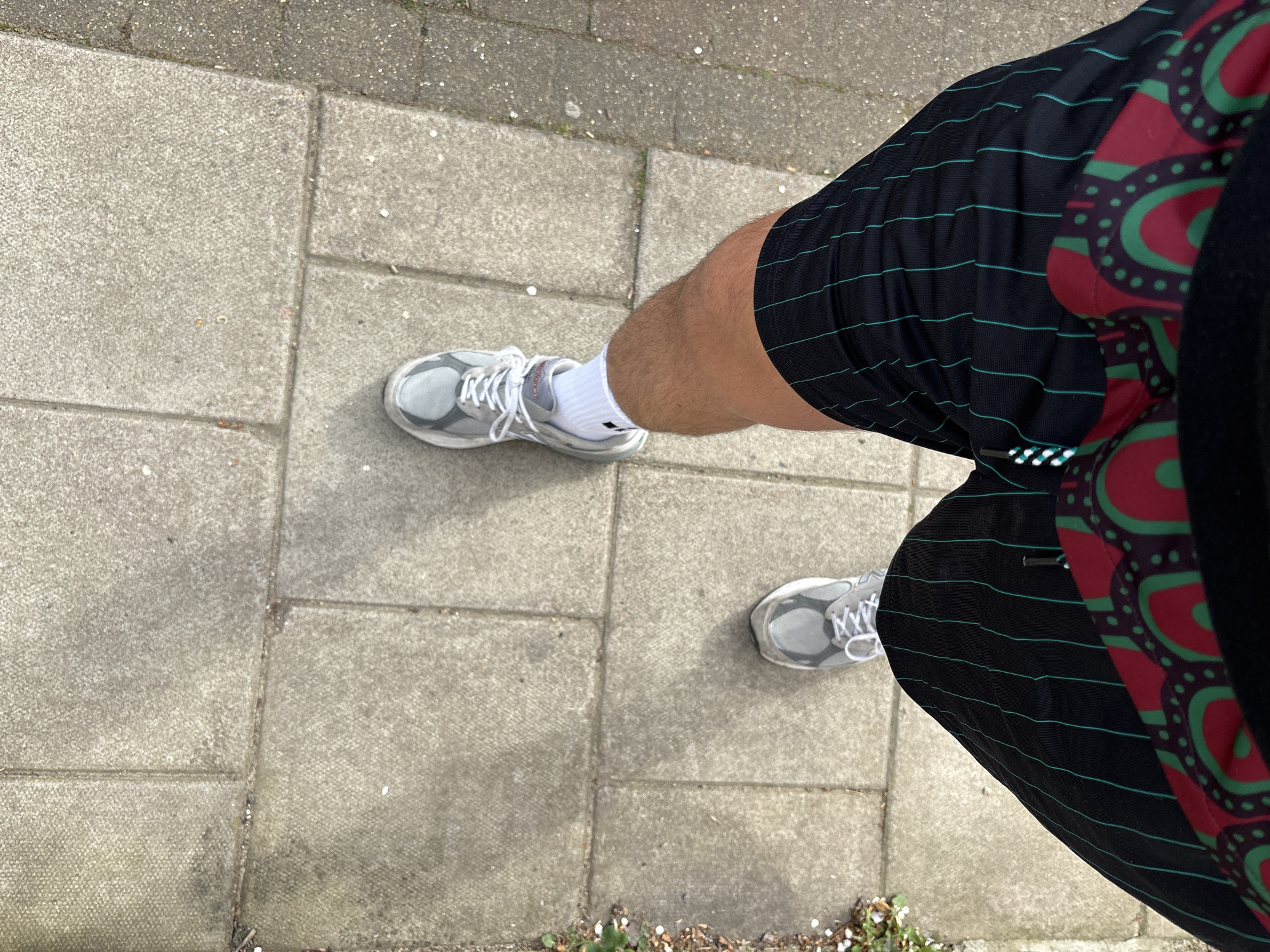 A person stands on a sidewalk paved with gray concrete rectangular blocks. They wear gray running shoes with white laces and white socks. Their shorts are black with thin teal horizontal stripes and a red and green patterned section along the side. The photo is taken from an overhead perspective. The location is indeterminate.