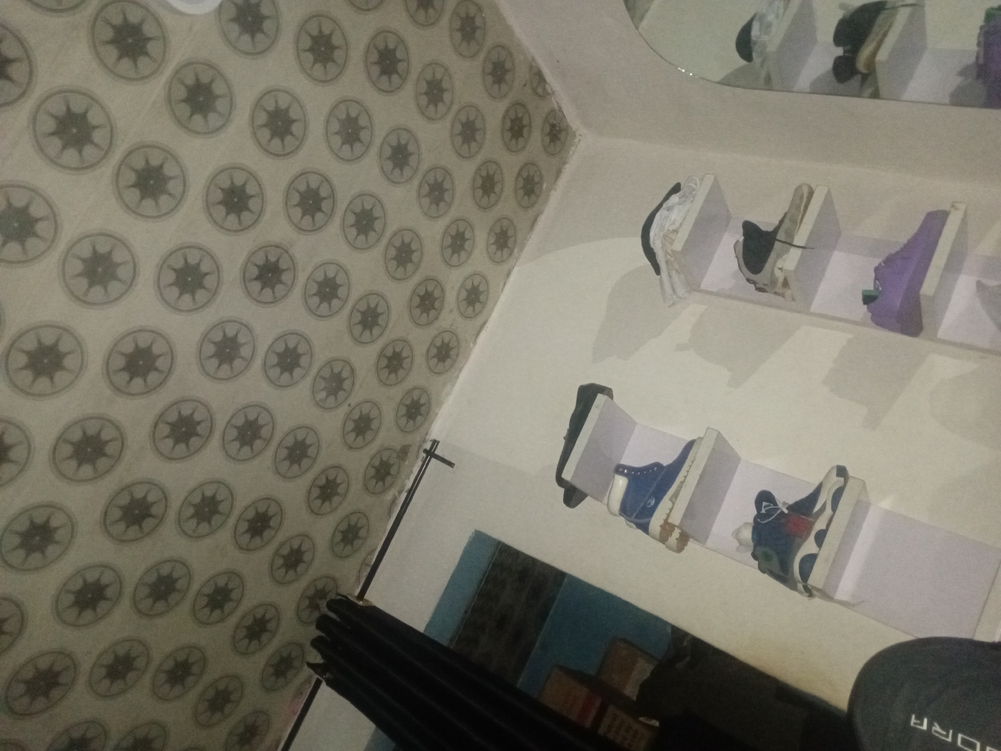 The image depicts an indoor scene in Ogbogbo, Nigeria, characterized by a distinct ceiling pattern and a display of footwear.
The ceiling is covered with a light-colored material, possibly plaster or tile, featuring a repetitive grid pattern of dark gray circular motifs. Each circle contains an eight-pointed star design. The pattern is consistent across the entire visible ceiling surface, extending into the corner where it meets a plain white wall.
The primary wall on the right is painted white and supports two vertical shelving units. Each unit consists of multiple rectangular white shelves.
On the left shelving unit, a blue high-top boot with a tan/white sole is placed on the top shelf. Below it, a dark blue, white, and possibly red high-top sneaker rests on the second shelf.
The right shelving unit displays several items. The top shelf contains a stack of white material, potentially clothing, topped by a dark fabric item. The second shelf holds a beige or off-white high-top sneaker with dark laces. Below this, a purple low-top sneaker is situated on the third shelf. The lowest visible shelf on this unit is partially obscured.
To the far right, a tall, arched mirror reflects portions of the shelving units and the shoes displayed on them.
On the left side of the image, a black curtain is drawn, covering a doorway or window opening. A black curtain rod extends horizontally above it. Through the opening, a light blue wall is visible in the background, with indistinct rectangular objects (possibly boxes or furniture) appearing in muted tones.
In the lower-left corner, the left shoulder and upper arm of a person are partially visible. The person is wearing a dark gray garment with the letters "ORA" visible, suggesting part of a brand name. The rest of the person is outside the frame. The overall lighting is dim, suggesting an indoor setting with either limited natural light or artificial illumination.