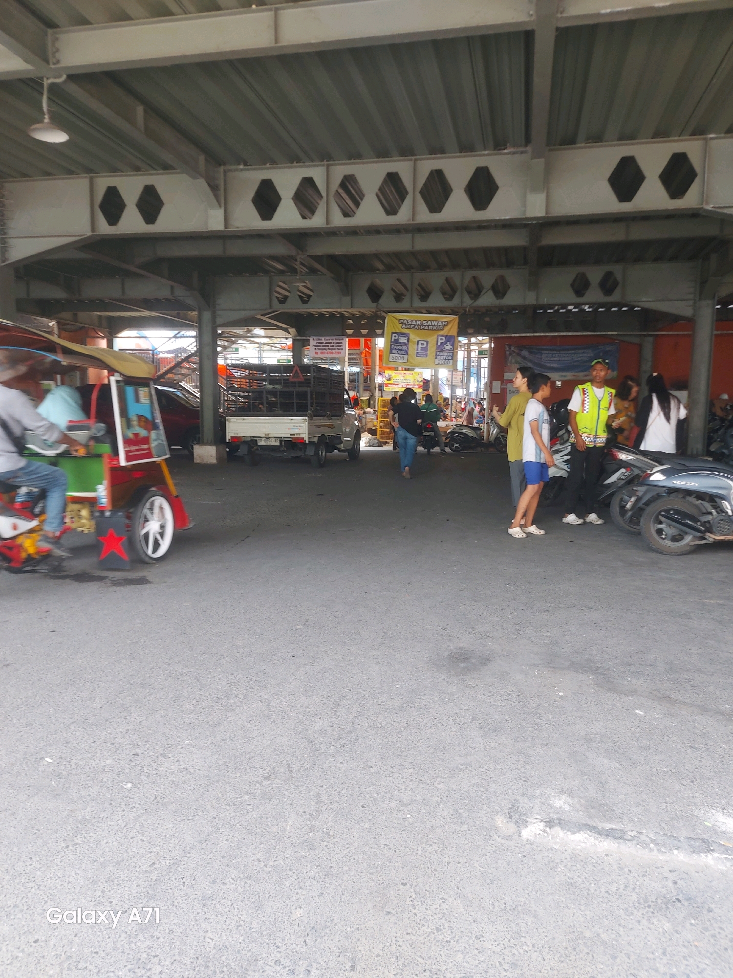 The image captures a scene under a covered structure, likely an overpass or a large canopy, in Makassar, Indonesia, possibly at a public market or transportation hub. The ground is paved with asphalt.
In the foreground on the left, a person is riding a colorful tricycle-like vehicle, possibly a becak, which is red and green with yellow accents and a yellow canopy. The vehicle has a prominent red star decal on its rear left wheel. The rider is wearing a grey long-sleeved shirt and blue jeans.
Further into the scene, a white utility truck with a metal cage at the back is parked. To the right of the truck, several people are gathered. Two young boys, one in a white t-shirt and blue shorts, and the other in a light green long-sleeved shirt, are standing together. A man in a yellow safety vest with reflective stripes and a green cap is standing nearby, appearing to be a worker or security personnel. A woman in a white long-sleeved shirt is also present. Several motorcycles are parked in the area.
In the background, banners and signs are visible, one of which reads "PASAR SAWAH AREA PARKIR" (Sawah Market Parking Area) with prices for "PARKIR MOBIL" (Car Parking) at 5000 and "PARKIR MOTOR" (Motorcycle Parking) at 2000. These signs suggest the location is a public area with parking facilities. The overall environment is utilitarian, with visible structural beams and corrugated metal roofing overhead.