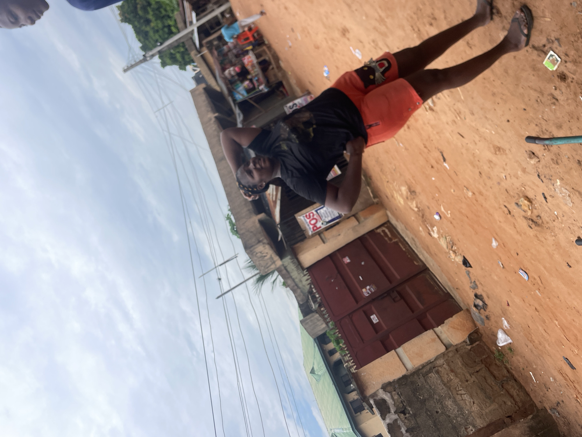 In Nkpor, Nigeria, a young woman with braided hair stands on a dirt road, posing with one hand on her hip and the other behind her head. She is wearing a black t-shirt and bright orange shorts. The background depicts a street scene with several buildings, including one with a green roof and a large red gate. A small market stall is visible to the right, with goods displayed and other people in the distance. Electrical wires are strung across the sky, indicating an urban or semi-urban setting. The ground is covered in reddish-brown dirt, with some scattered debris, typical of the environment.