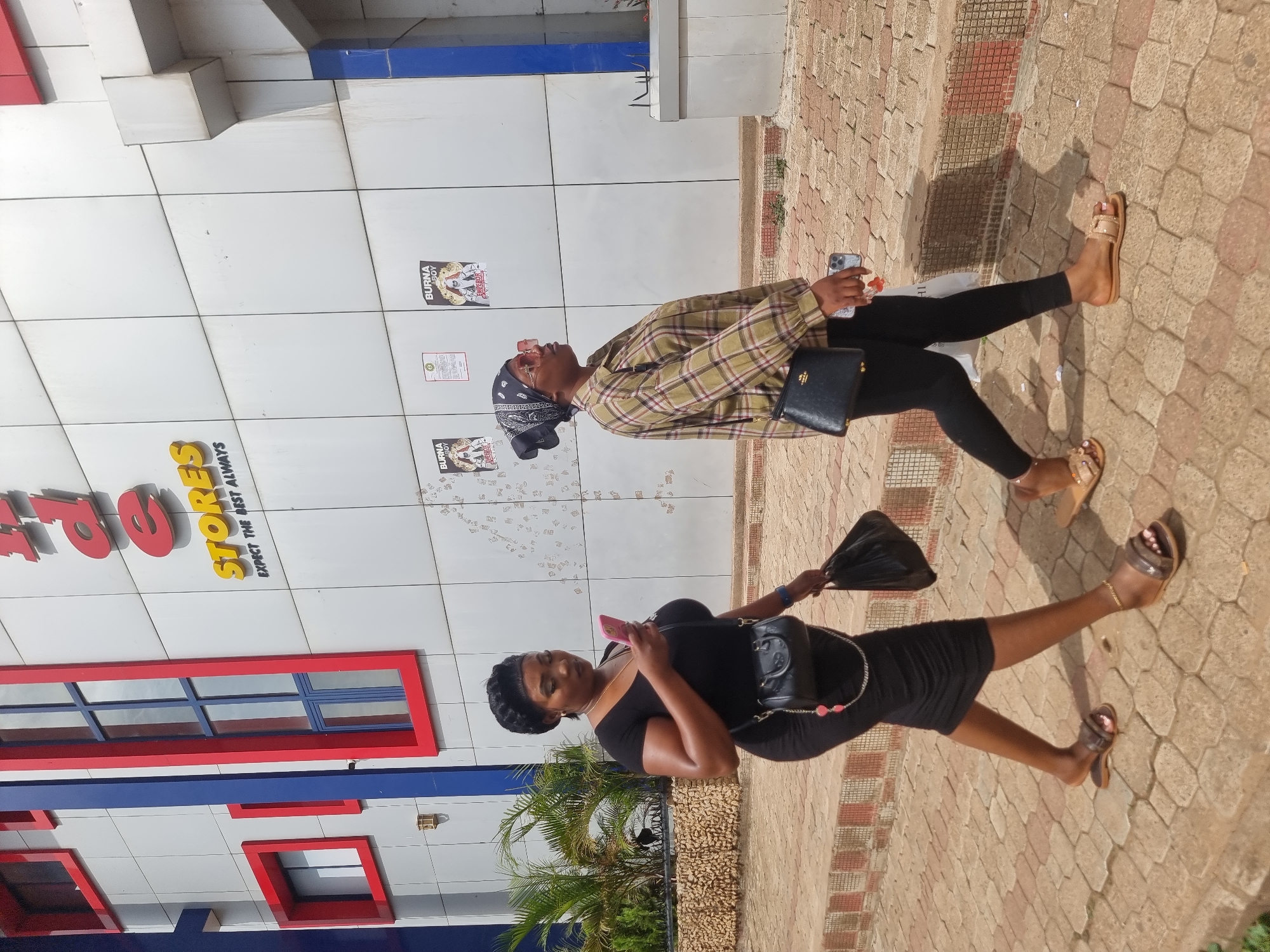 Two women are walking on a brick-paved sidewalk next to a building in Jos, Nigeria. The building has a sign that says "STORES" with the tagline "EXPECT THE BEST ALWAYS". The building is white with blue and red trim. One woman is wearing a black dress and holding a pink phone and black handbag. The other woman is wearing a plaid shirt, black pants, and a bandana, and she is holding a phone and a black Prada handbag. There are posters on the wall behind the second woman. A small palm tree is near the first woman.