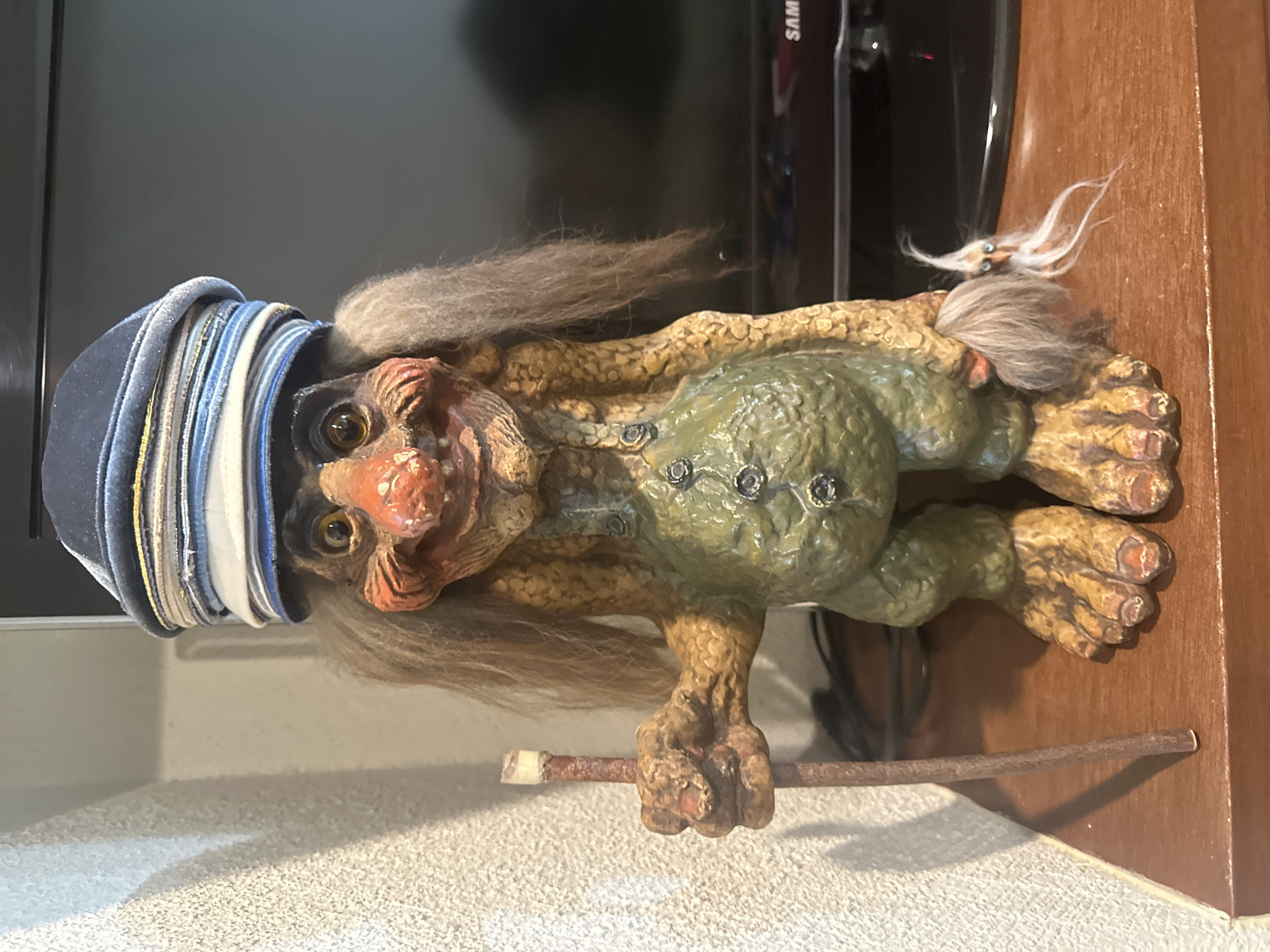 A troll doll with grey hair wearing a blue and white striped hat and holding a brown stick is sitting on a wooden surface. The doll is green and tan with a large nose and feet. A black television sits behind the doll. The image was taken indoors. The location is not evident in the image.