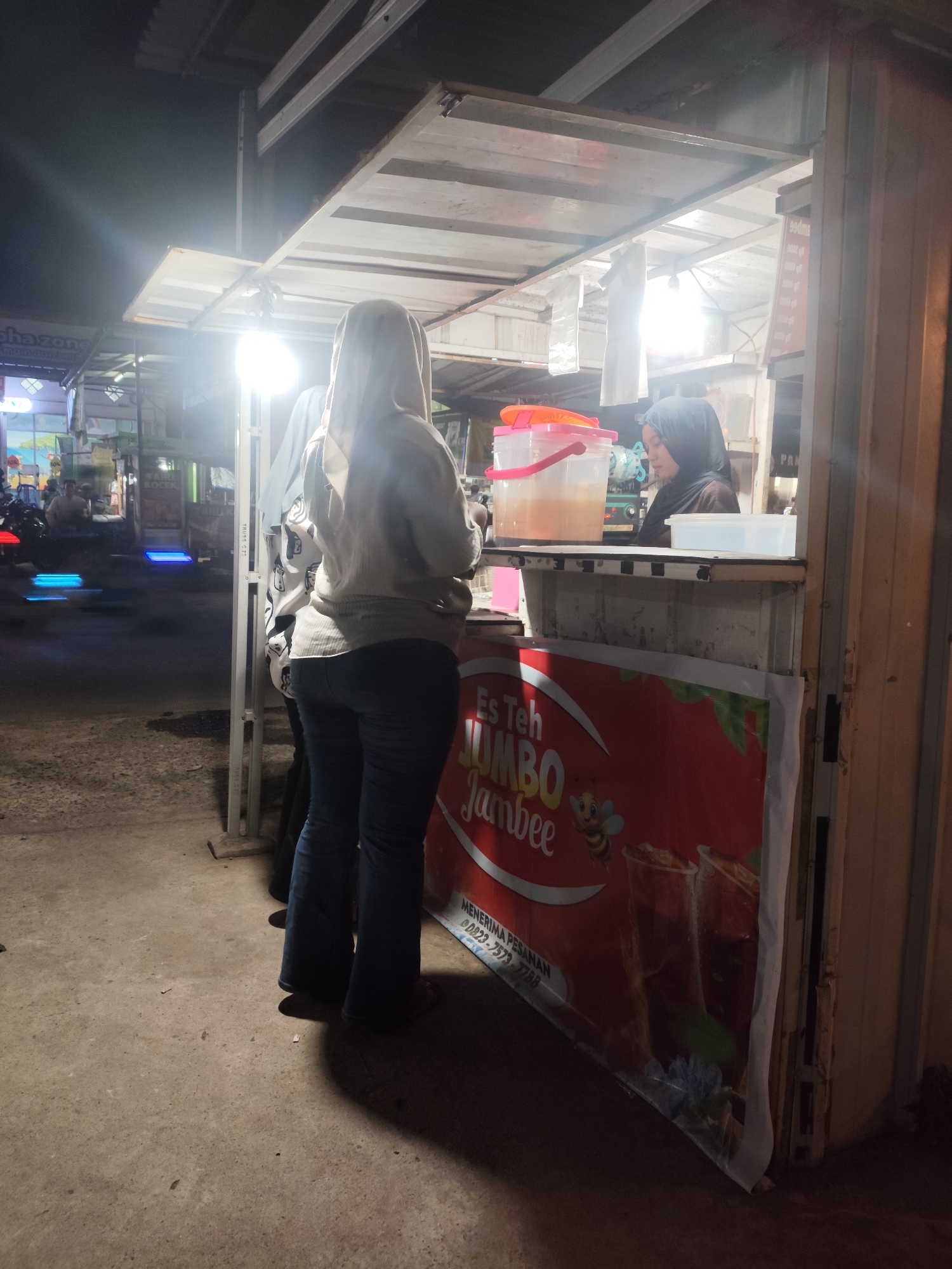 The image captures an outdoor nighttime scene in Palembang, Indonesia, likely at a street-side food or drink stall. In the foreground, two individuals are visible. One person, viewed from behind, is wearing a light-colored hooded jacket and dark, bell-bottom jeans. They are standing with their back to the camera, facing a stall. To their right, and slightly behind them, is another person wearing a dark hijab and a dark top, who appears to be serving at the stall.
The stall itself has a counter with several items on it, including large plastic containers of beverages, one with an orange lid containing a yellowish liquid, and a white plastic container. Behind the counter, there are more containers and supplies. A prominent red banner with white text and imagery of drinks is displayed on the front of the stall. The banner appears to advertise "Es Teh Jumbo Jambee" and includes a small image of a bee. Text below this reads "MENERIMA PESANAN" and a phone number.
The environment suggests a busy street, with illuminated signs and the blurred motion of vehicles in the background, indicated by streaks of blue light. The ground is paved and appears slightly damp or wet. The lighting is harsh, with a bright light source illuminating the area directly above and in front of the stall. The overall impression is one of a typical roadside commercial activity at night in an urban Indonesian setting.
