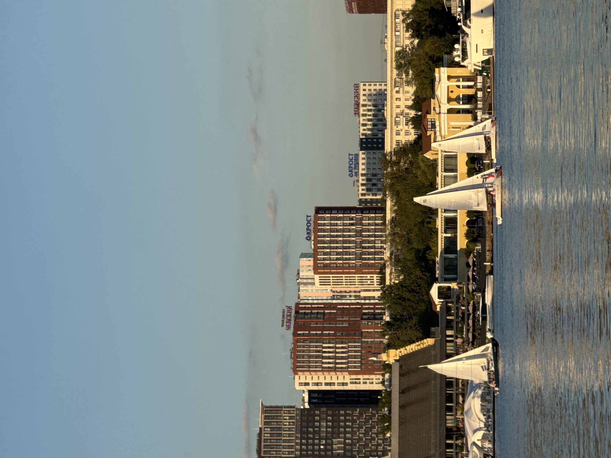The image depicts a sunny day over the city of Khimki, Russia, with a body of water in the foreground and a cityscape in the background. Several sailboats are visible on the water, with one prominent sailboat in the left-center of the frame, having two people on board. To the right, two more sailboats are sailing, with a large white yacht positioned further to the right. The shoreline features a building with a yellow facade, flanked by trees and other structures. Above the shoreline, several modern buildings rise into the clear blue sky, some with visible signage including "НЕВСКИЙ" and "АКРОСТ." The overall scene suggests a recreational activity on the water within an urban setting.