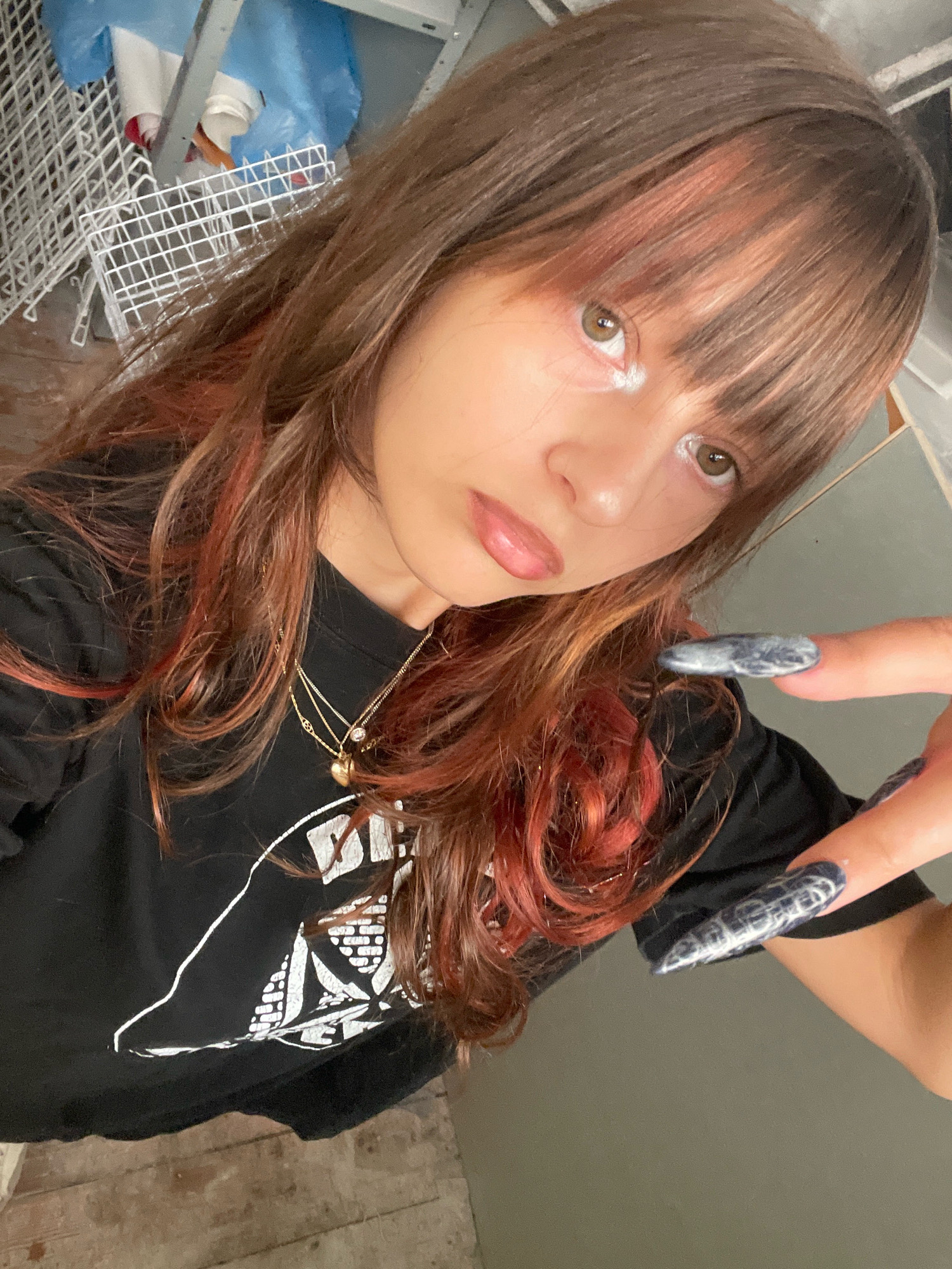 A young woman with long, brown hair streaked with red is taking a selfie. She is wearing a black t-shirt with a white graphic design and a gold necklace. Her nails are long and painted with a dark, marbled design. She is looking directly at the camera with a neutral expression. The background appears to be an indoor space, possibly a garage or storage area, with shelves, white wire racks, and blue plastic sheeting visible. The floor is made of wooden planks. There is no indication of this location being in Raanana, Israel.