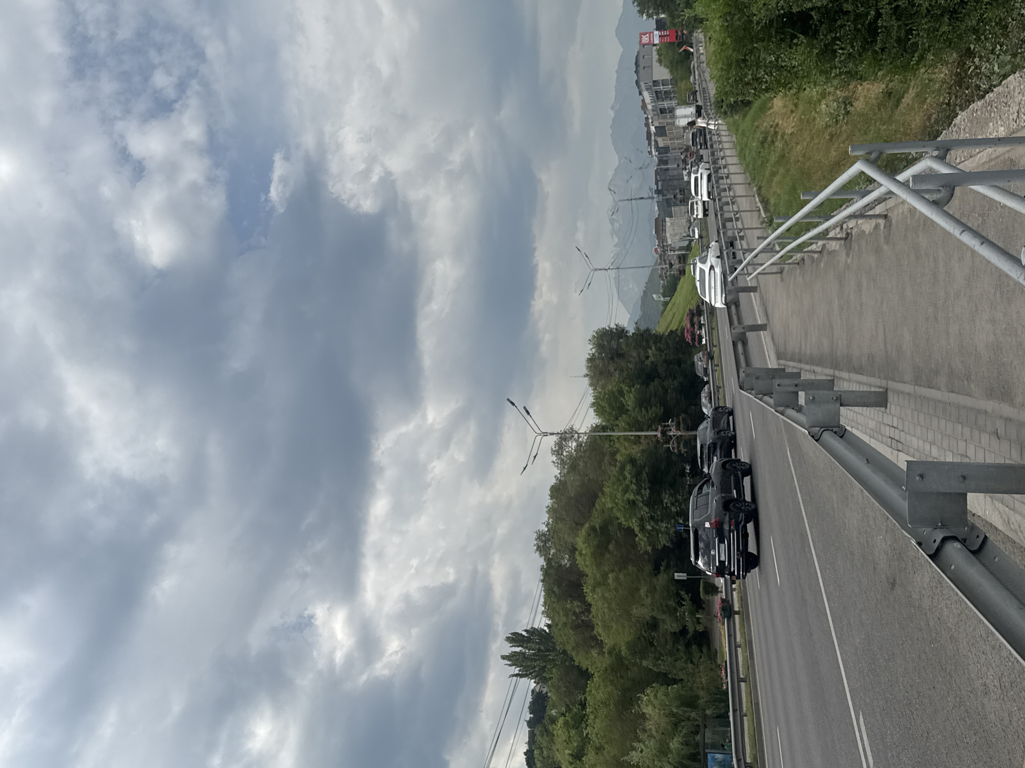 The image shows a view of a multi-lane road with vehicles in Almaty, Kazakhstan, under a cloudy sky. On the right side of the road, there is a pedestrian walkway separated by a metal railing. Further to the right, a grassy slope covered in green bushes rises. In the background, there are buildings and some distant snow-capped mountains. The road appears to be in a city or suburban area. The time of day is daytime, likely afternoon, given the cloud cover and light.