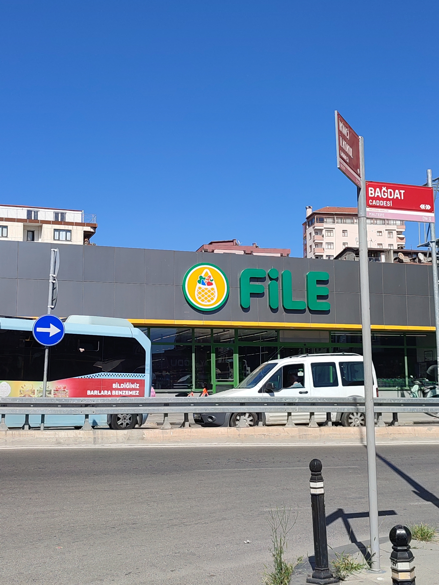 This is a medium shot, taken from a low angle on a sunny day, of the front of a "FILE" supermarket in Maltepe, Turkey. A bright blue sky fills the top half of the frame. The supermarket has a grey facade with large green "FILE" lettering. To the left of the lettering is a circular green and yellow logo featuring a pineapple. Below the logo, on the side of a blue bus, is a red advertisement with white text that reads "BİLDİĞİNİZ BARLARA BENZEMEZ". A white van is parked in front of the supermarket.
A street sign in the upper right corner displays "BAĞDAT CADDESİ" and "MALTEPE" in red lettering against a red background, indicating the location. A thin grey pole stands in the foreground, casting a shadow. Several bollards, one black and one grey, are also visible on the sidewalk. The supermarket is situated across a road with a metal barrier separating it from the sidewalk. The background features the tops of residential buildings.