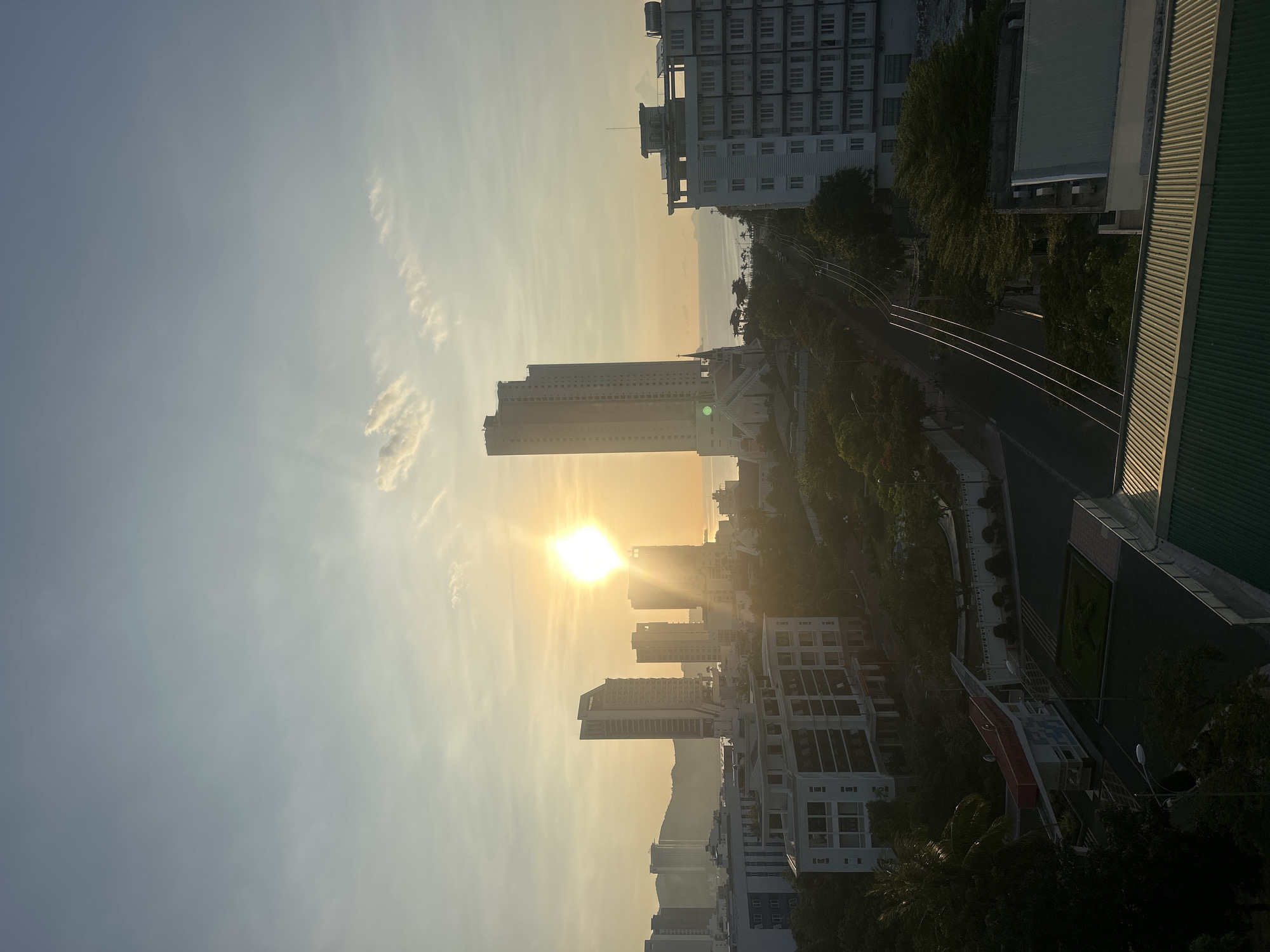 The image depicts a sunrise over Bac Nha Trang Ward, Vietnam. The sun, a bright orb, is positioned behind a tall, slender building, radiating light. Silhouetted buildings of varying heights dot the cityscape. Vegetation is also visible interspersed among the buildings, lining a road. The sky is a gradient of blues and yellows, with wisps of clouds scattered across it.