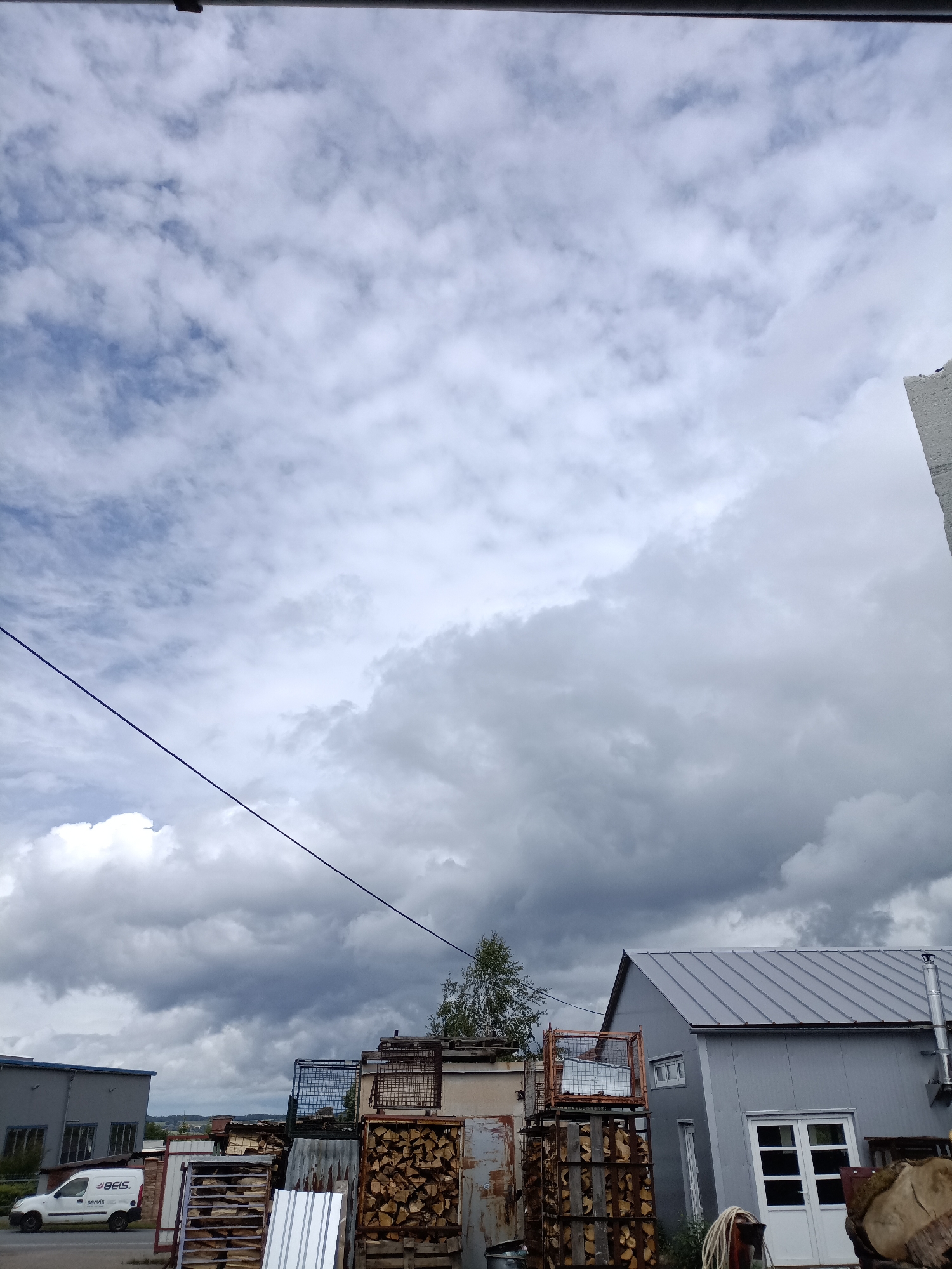 This image depicts an industrial or storage area in Dalovy, Czechia, under a predominantly cloudy sky. The sky is characterized by a mix of fluffy white clouds and darker, heavier grey clouds, with glimpses of lighter blue in the upper left, suggesting an overcast day with dynamic weather. A single dark blue utility line stretches diagonally across the sky from the top left to the lower center.
In the midground, several structures are visible. On the left, a grey, boxy building with rectangular windows and a flat roof stands. In front of it, a white commercial van is parked, featuring the text "BELS" prominently in its logo, with "servis" written underneath. To the right, another grey building with a sloped, corrugated metal roof, a white double door, and several windows is present. Between these buildings, a tall, slender tree with sparse green leaves is visible.
The foreground and midground are densely packed with various industrial and raw materials. Numerous stacks of chopped firewood are neatly arranged within sturdy metal crates or cages. Alongside these, there are piles of wooden pallets, corrugated metal sheets, and other miscellaneous industrial debris, suggesting a busy storage or processing facility. A large section of a tree trunk or log is visible on the far right. No people are present, and the scene conveys a functional, utilitarian atmosphere during what appears to be daytime.