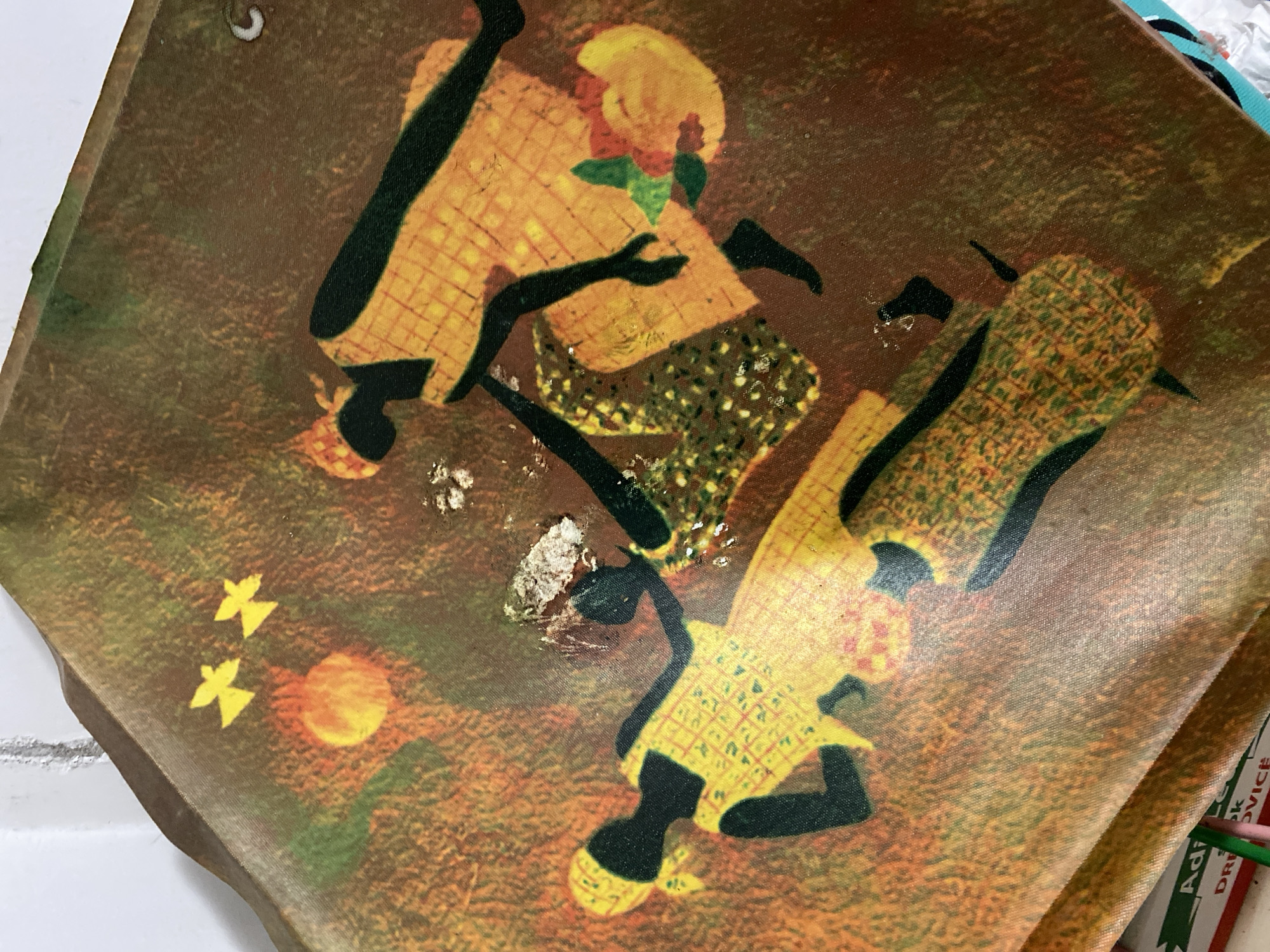 The image shows a rectangular tray with an African-themed design. The tray is brown and features stylized figures of people with black limbs and heads, and yellow patterned clothing. Two of the figures appear to be dancing, while a third figure is in the upper portion of the tray. A faded yellow sun and stylized birds are visible in the lower left corner. There is some damage to the surface of the tray in the form of a silvery substance. The tray is set on a white surface and partially obscures a green and white object with the text "Adi..." and "...ovica". The image does not contain any information regarding the location of Budva, Montenegro.