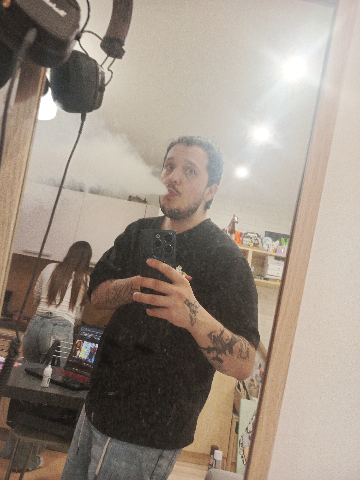 A man is taking a mirror selfie indoors. He is exhaling vapor, holding a black smartphone with three camera lenses. The man wears a black t-shirt and light blue jeans. He has tattoos on both arms. A woman is standing in the background, facing away from the camera, wearing a white long-sleeve shirt and blue jeans. A black desk with a laptop and a small bottle on it is visible. Overhead headphones are hanging in the upper left corner. A light-colored cabinet and shelf are behind the woman. Based on the decor (especially the brick wall) and the man's social media profile, the location appears to be in Penza, Russia.