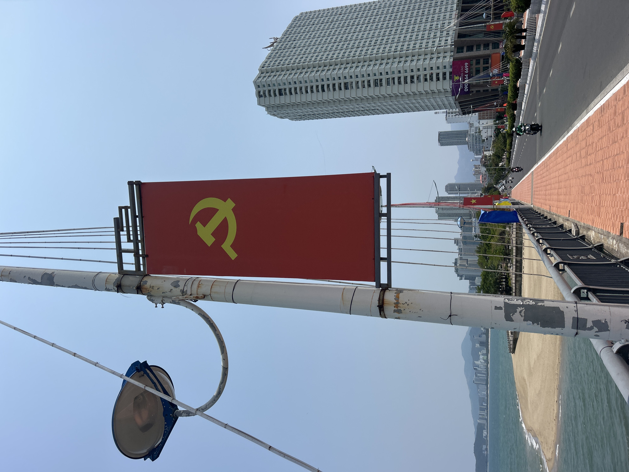 The image shows a red flag with a yellow hammer and sickle symbol, a symbol often associated with communism, prominently displayed. The flag is mounted on a light pole with a street lamp attached, situated on a bridge. In the background, there is a cityscape with a tall, modern skyscraper. A roadway with pedestrian walkway is visible on the right side of the image with several motorbikes are traveling in the same direction. A beach is also visible below and to the right of the bridge. Based on the background scenery, the location is likely Nha Trang, Vietnam.