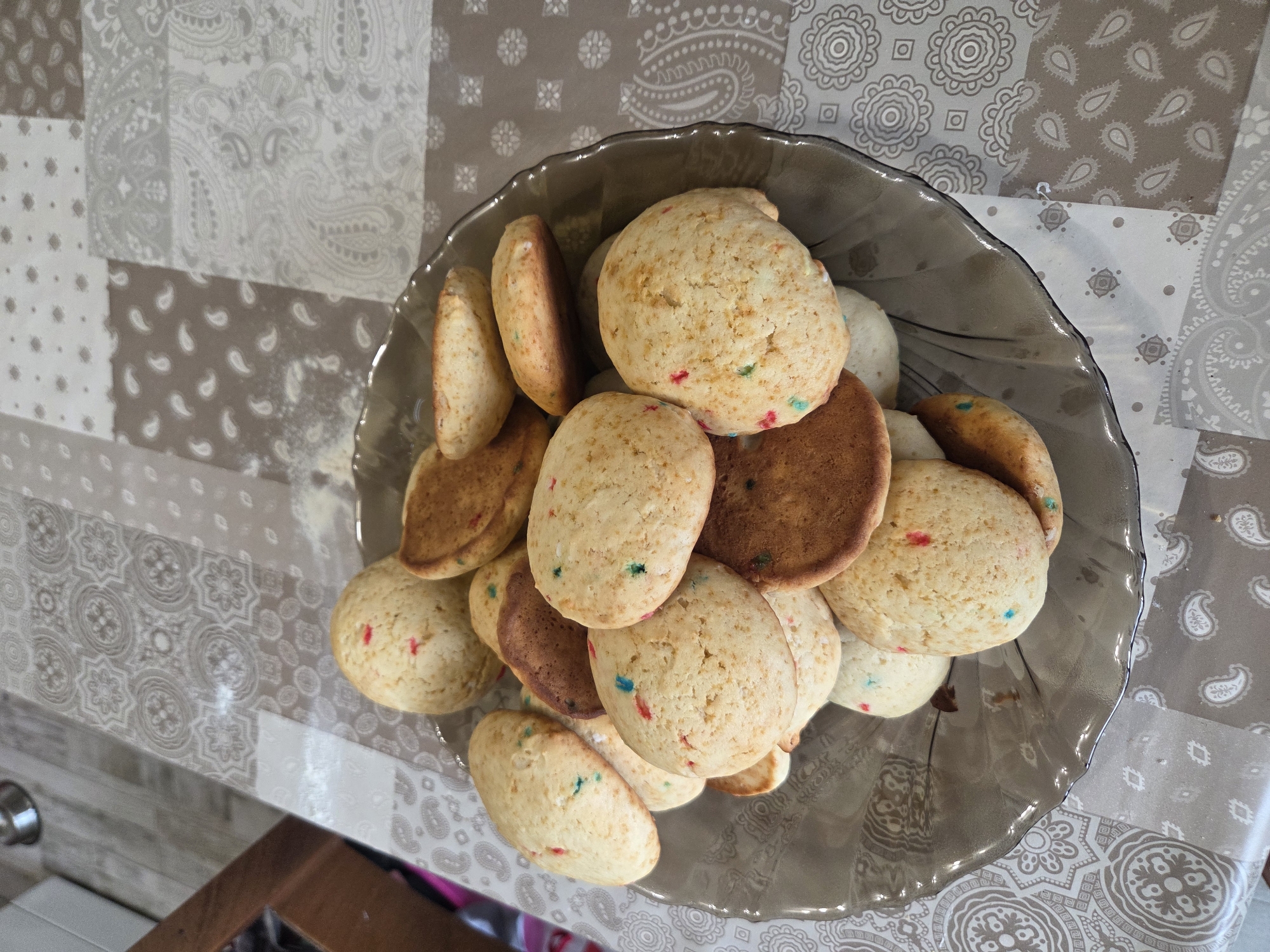 A bowl of colorful cookies sits on a table in Plovdiv, Bulgaria. The table is covered with a patchwork tablecloth. The cookies are round and appear to be sugar cookies with colorful sprinkles. Some are slightly browned. They are piled in a gray glass bowl. The table is next to a counter.
