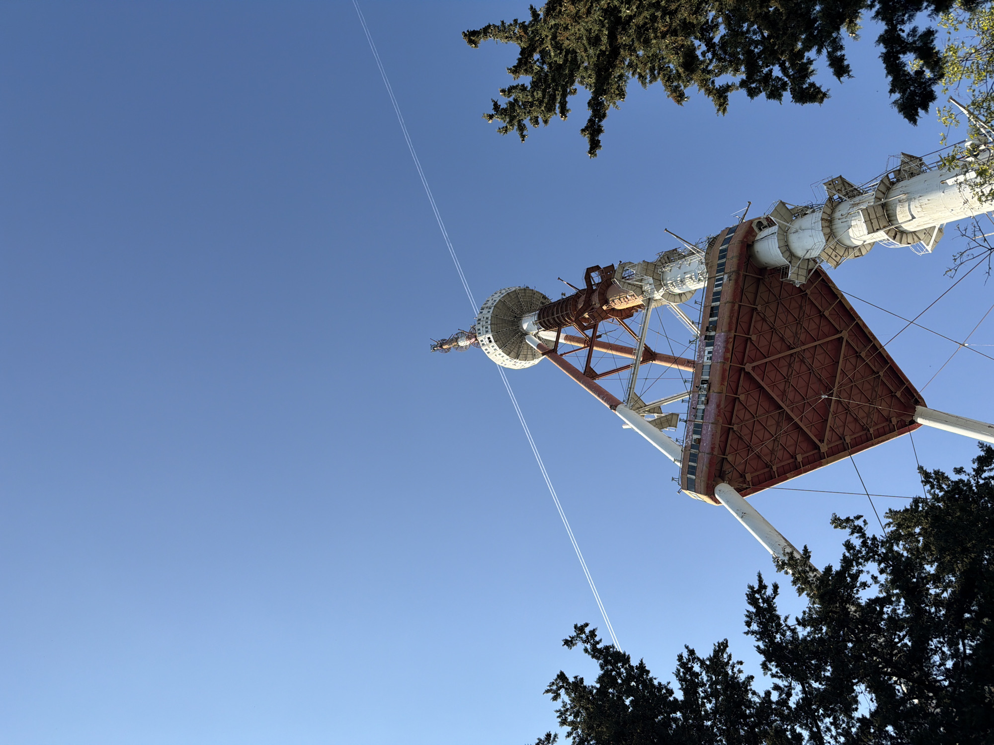 This low-angle, wide shot captures the imposing Tbilisi TV Tower against a vast, clear blue sky, viewed from its base. The prominent structure, a distinctive landmark in Tbilisi, Georgia, features a red-brown latticework base transitioning into white cylindrical sections adorned with various communication antennae and platforms. The perspective emphasizes the tower's immense height, stretching diagonally across the frame from the bottom right towards the upper left.
Dark green foliage from evergreen trees, possibly conifers, frames parts of the tower, appearing in the top right and bottom right corners of the image. The bright, cloudless sky indicates a sunny day, likely morning or afternoon, given the clear illumination on the tower's surfaces. A notable detail in the sky is the presence of two distinct, parallel white contrails, left by an aircraft, arcing diagonally across the upper left portion of the blue expanse. The overall impression is one of architectural grandeur set against a serene natural backdrop. There is no visible text in the image.