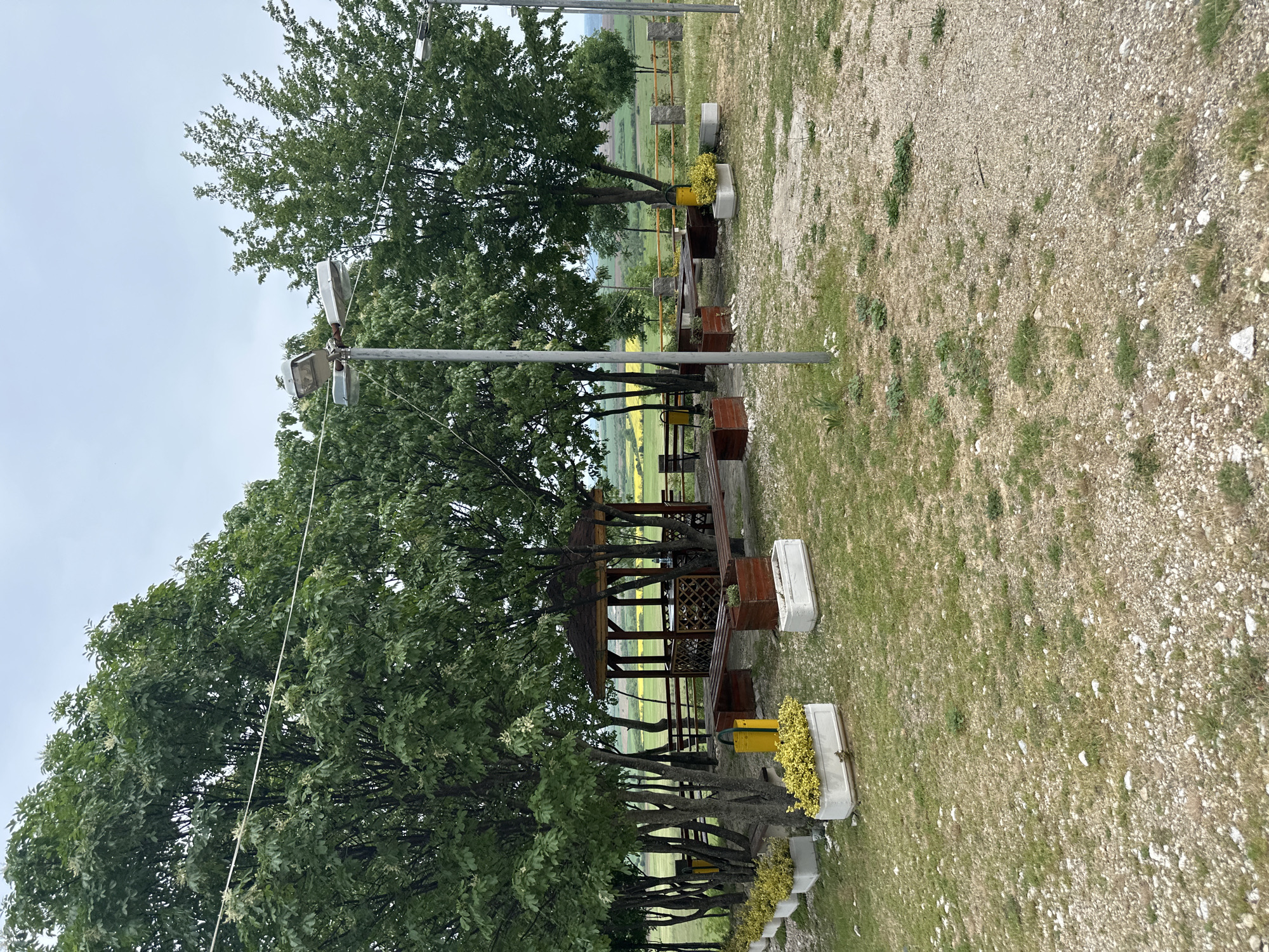 The image depicts an outdoor setting near Gara Oreshets, Bulgaria. The foreground consists of gravel and grassy areas interspersed with occasional vegetation. Several trees with green foliage line the area, partially obscuring a wooden gazebo-like structure with benches. Planter boxes are situated near the trees, some containing yellow flowers. Above the trees are lampposts. In the background, a landscape of fields and distant hills can be seen under a cloudy sky.