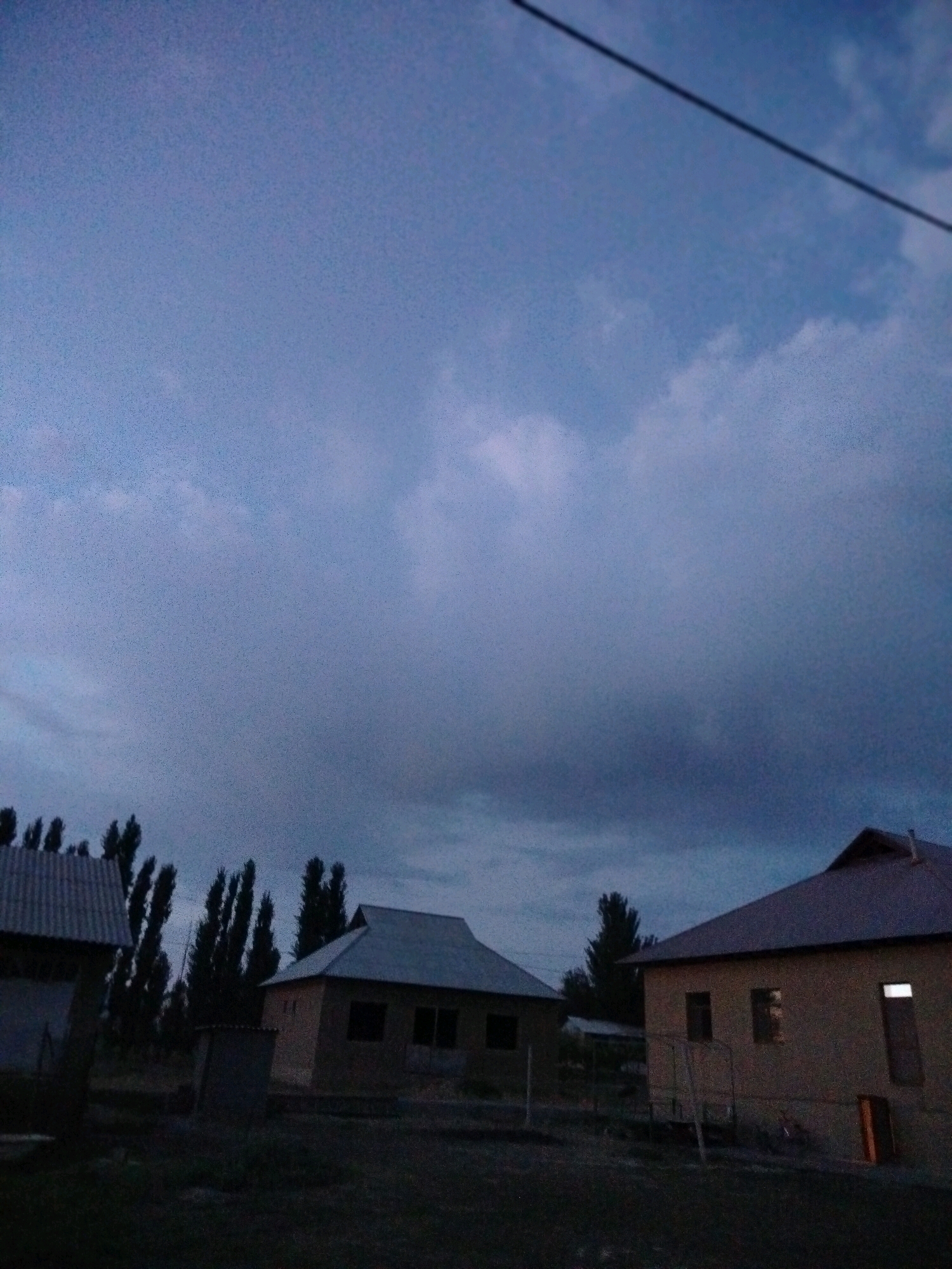 The image depicts a rural scene, likely on the outskirts of the city of Көлкент, Kazakhstan, as indicated by the context. The sky is overcast with a mix of light and dark clouds, suggesting either dawn, dusk, or a stormy day. In the foreground, two modest, single-story houses are visible. The house on the right has a dark roof and cream-colored walls, with several windows and a door. One window on the right side of this house is illuminated from within. The house on the left also has a dark, corrugated roof and appears to be a simpler structure. Tall, slender trees are scattered throughout the background, silhouetted against the sky. The ground in front of the houses is dark and appears to be unpaved. There are no people visible in the image. A power line cuts diagonally across the upper portion of the sky. The overall impression is one of a quiet, undeveloped residential area.