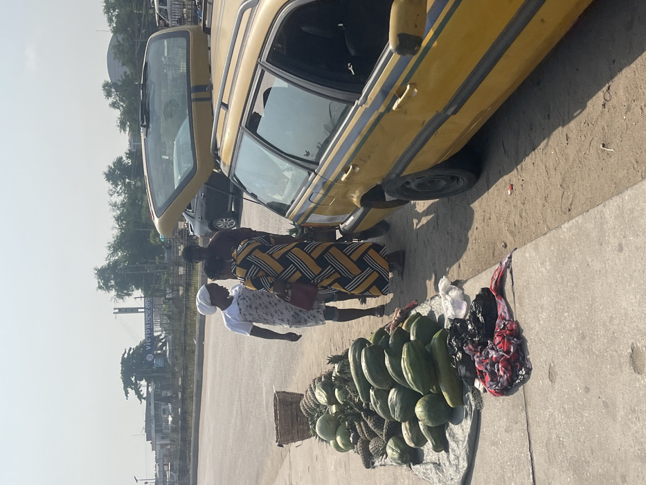 The scene depicts a roadside market setting in Ikota, Lagos, Nigeria, under bright daylight conditions. A blue sign reading "VICTORIA ISLAND" is visible in the background. On the left, a large display of fruits, including numerous green papayas, pineapples, and spherical green fruits (possibly watermelons or gourds), rests on a grey tarp on the ground, adjacent to a concrete curb. A woven basket is positioned near the top of the fruit pile. Below the fruit display, black plastic bags and crumpled red-and-white patterned fabric are situated on the concrete curb.
Three individuals are gathered to the right of the fruit stall. A woman wearing a white head covering, a white short-sleeved top, and a light-colored patterned wrap stands facing right, seemingly in conversation. She faces a second woman, whose back is largely to the camera, wearing a long dress with yellow, black, and white geometric patterns and carrying a small reddish-brown handbag. A man in a dark top is partially visible behind the second woman. To the far right, a yellow station wagon, featuring blue stripes along its side, is parked on a dirt shoulder with its rear hatch open, exposing the cargo area. A paved road stretches behind the individuals and the fruit display, with distant buildings, trees, and other vehicles visible under a hazy sky.