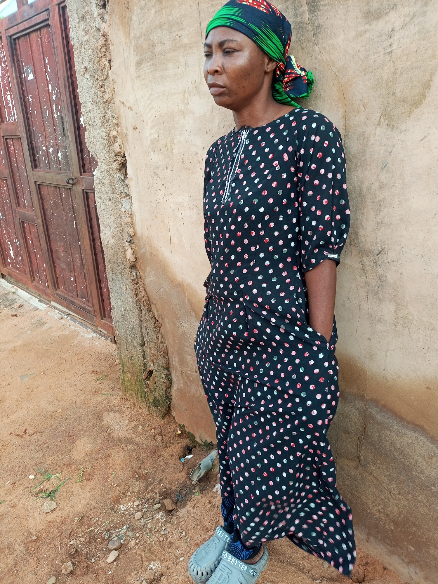 A woman stands with her back to a wall, her body angled away from the camera. She is wearing a black dress with colorful polka dots and a green, red, and black headscarf. Her right hand is in her pocket. The wall behind her is a rough, textured beige surface. To her left is a large, weathered wooden gate with peeling red paint. The ground in front of her is dry, reddish-brown dirt with scattered small rocks and some sparse green vegetation. The scene appears to be an outdoor setting in an urban or semi-urban environment, possibly in Tempure, Nigeria, given the context.