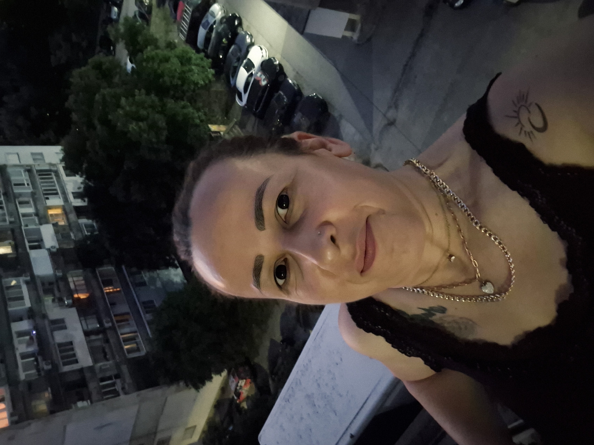 A selfie taken at dusk on a balcony overlooks a city street lined with parked cars. A woman in a black lace-trimmed top is in the foreground, facing the camera. She wears multiple gold necklaces, one with a heart-shaped pendant, and has a tattoo of a crescent moon and sun on her left shoulder. Behind her, a large apartment building with lit windows and trees partially obscure the view of a parking lot filled with various cars. The lighting suggests late evening or early night.