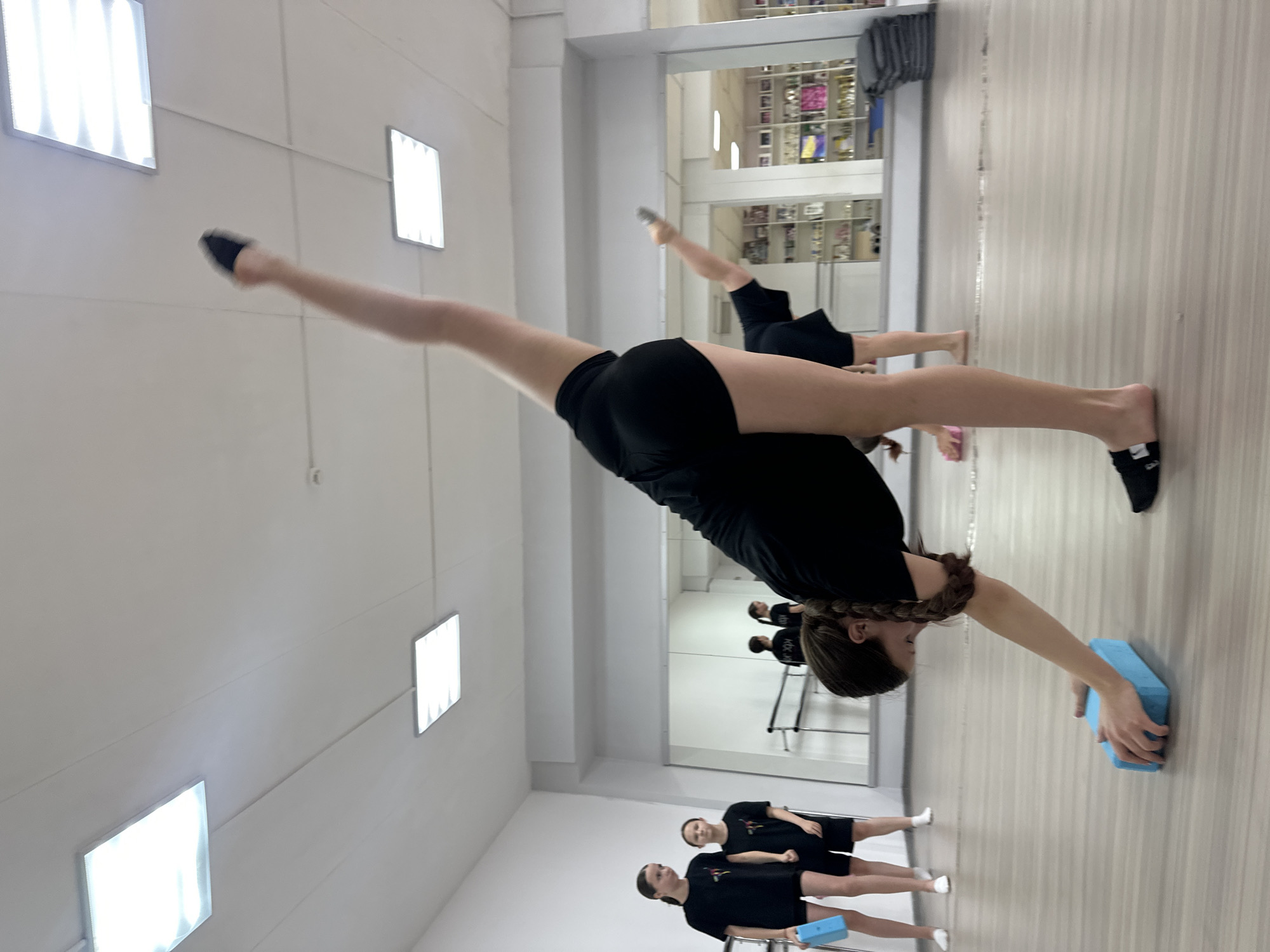In a dance studio with a light-colored wooden floor and white walls, four young females are practicing. Two are in the foreground, with one in black shorts and a black t-shirt in a split, her right leg extended upwards and her left hand touching a blue yoga block on the floor. Her hair is in a braid. The other in the foreground is facing away from the camera and is in a split with her right leg extended upwards. Two other young females are in the background, also wearing black t-shirts and black shorts, standing near a mirror. The studio has large ceiling lights and a ballet barre along the wall, with shelves filled with trophies and awards in the background.