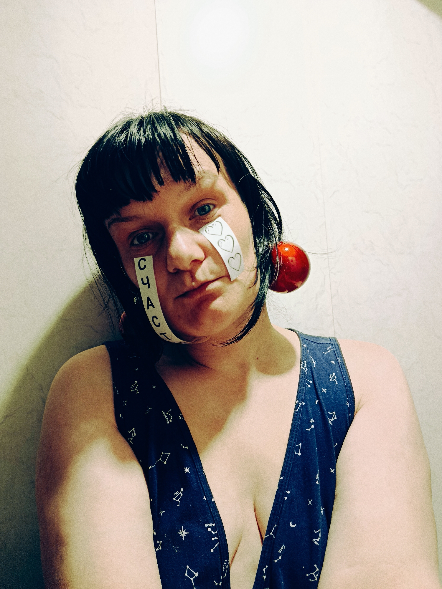 A person with short black hair and bangs poses in front of a light-colored wall. They are wearing a navy blue tank top with white constellation patterns. A strip of white tape with the word "СЧАСТЬ" written vertically in Cyrillic is stuck to their left cheek, while another strip of tape with drawn hearts is on their right cheek. A large red ball-shaped object is attached to their right ear. The location is unknown.