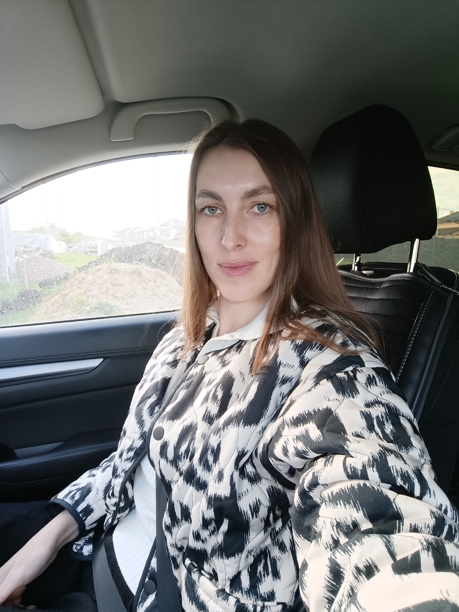 A woman is seated in a car, facing the camera. She has long brown hair and blue eyes. She is wearing a white shirt under a black and white patterned jacket, and a black seatbelt. The car has black leather seats. Outside the car window is a view of what appears to be a construction site with piles of dirt and some partially constructed buildings. Based on the metadata, the location is identified as Chaikovsky, Russia.