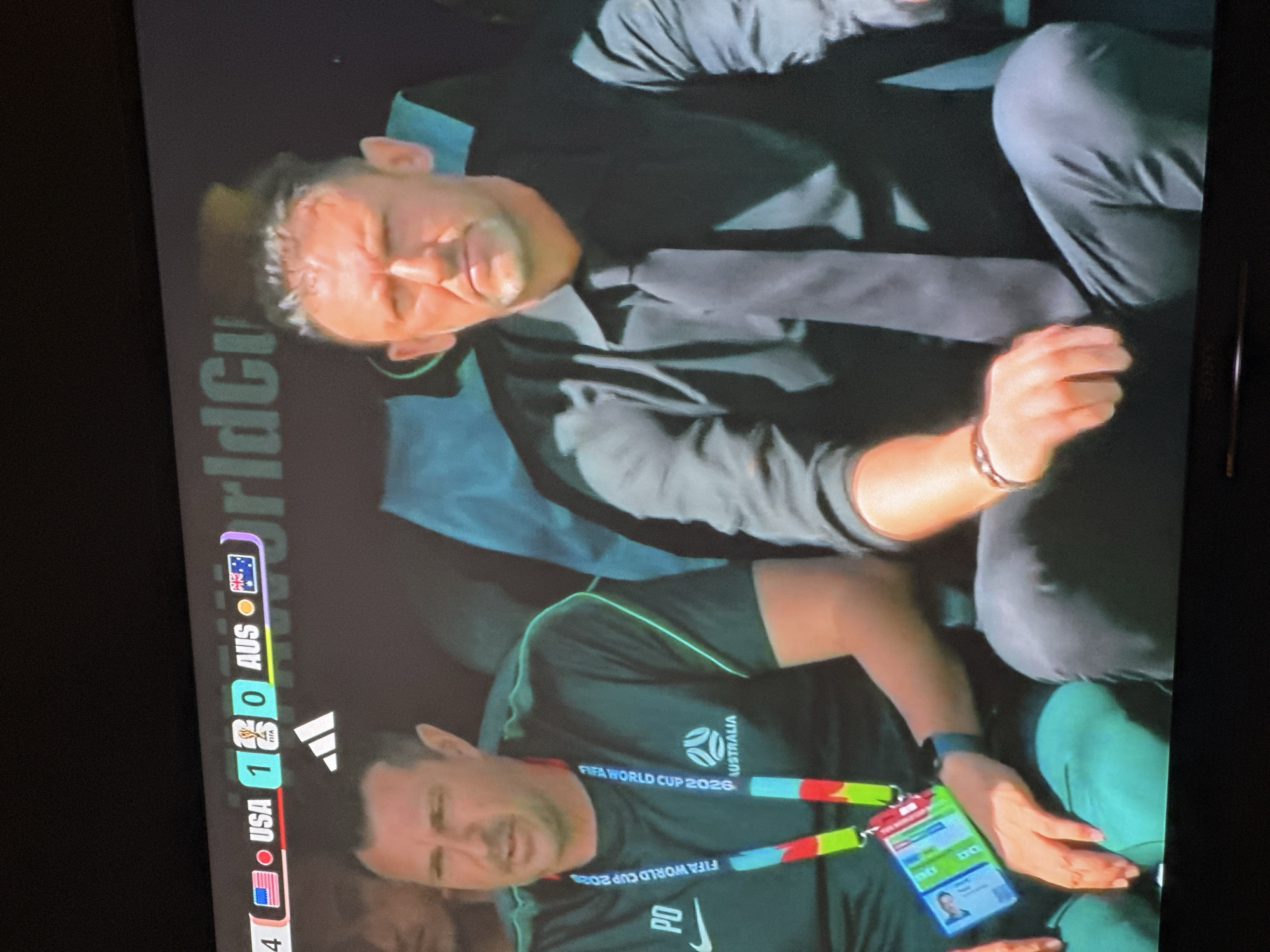The image shows a close-up of two men sitting on a bench, presumably in a stadium. A scoreboard in the background indicates a soccer match between USA and AUS, with USA leading 1-0. The man on the left is wearing a green t-shirt with "PO" on it and a lanyard with multiple badges, one of which has the FIFA World Cup 2026 logo. He appears to be speaking. The man on the right is wearing a black dress shirt and tie and has his hair pulled back. He is looking forward with a serious expression. The environment appears to be indoors, with artificial lighting. The provided location "Gura Top, Nigeria" is not visible in the image.