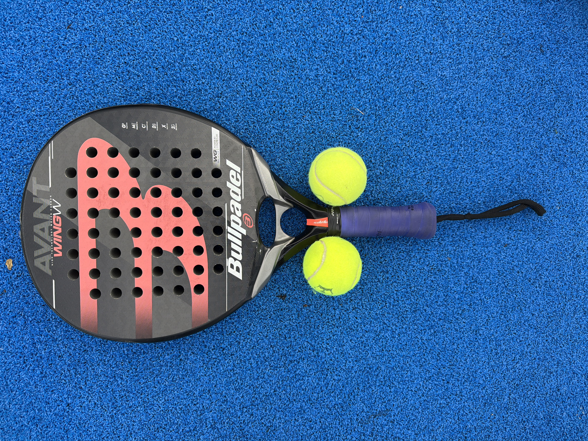 A padel racket with the brand "Bullpadel" and model "Avant WingW" is laid out on a textured blue surface, resembling artificial turf. Two yellow tennis balls are positioned on either side of the racket's handle, resembling eyes. The racket itself is black and red, with circular holes on its hitting surface and the brand logo prominent in the center. The handle is wrapped in purple grip tape, and a black wrist strap hangs from the bottom. The blue surface appears to be an outdoor sports court, possibly a tennis or padel court. The scene is devoid of any people. The location is identified as Vaterstetten, Germany.