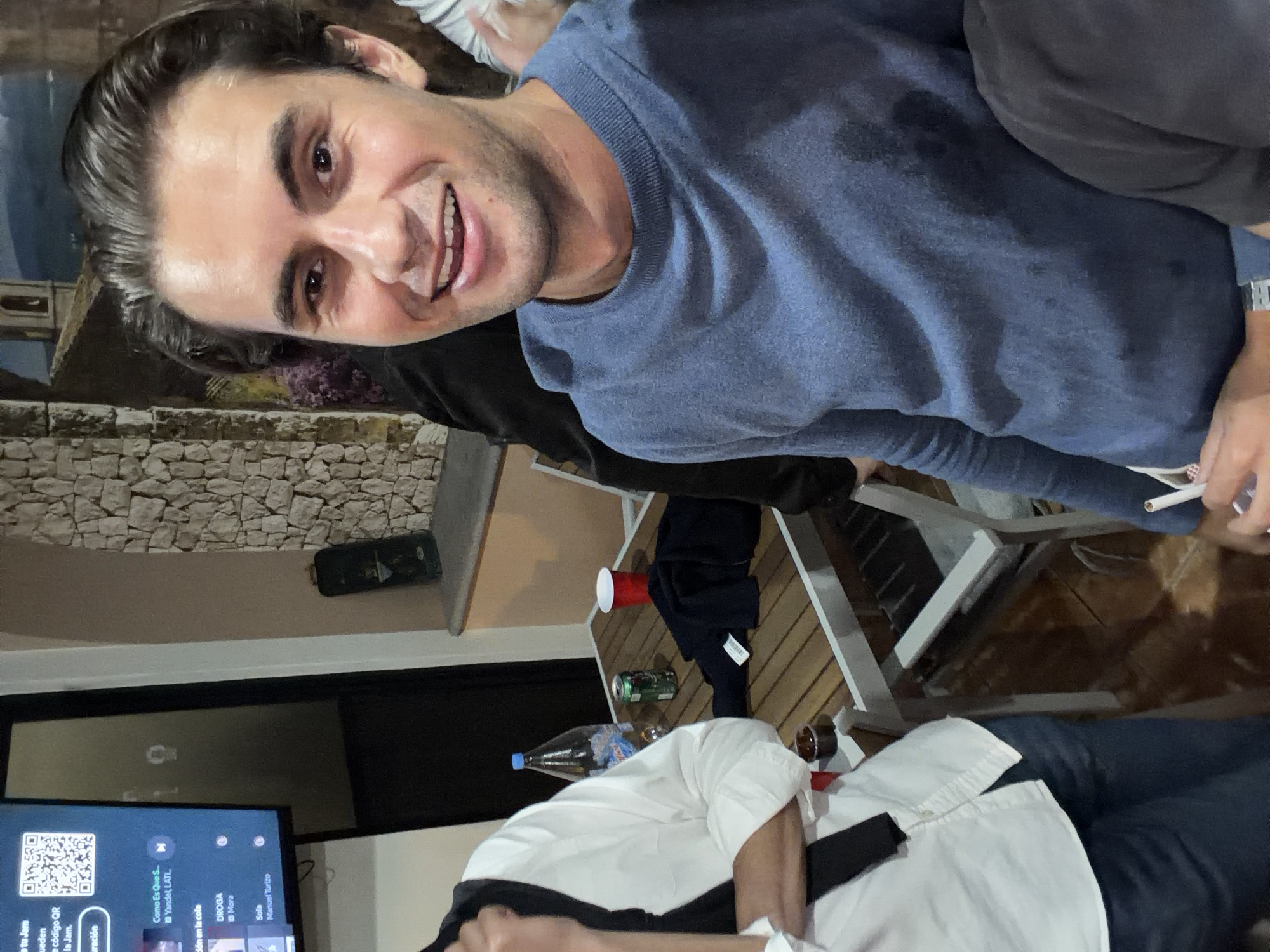 A candid, close-up image shows a man smiling into the camera, seated with others at an outdoor table. The man is wearing a blue sweater and holds a cigarette. Part of another person is visible, wearing a white shirt and black pants. A TV screen is in the background, along with a stone-textured wall. On the wooden table are several items including a red cup, a green can, a bottle of water, and what appears to be a brown liquid in a glass or container. The location is San Pedro Cholula, Mexico.