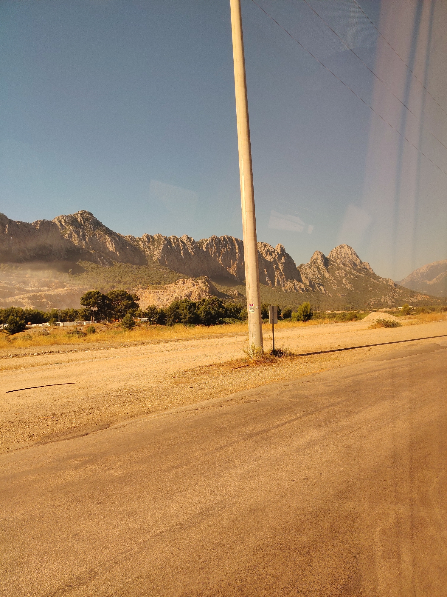 A wide shot captures a dry, arid landscape with a rough asphalt road in the foreground. The road curves slightly to the right and appears to be a dirt road mixed with asphalt. To the left of the road, a sandy area stretches out. In the middle ground, a row of trees stands out against the backdrop of rugged, rocky mountains. The mountains are jagged and appear to be covered in sparse vegetation. A single, tall, light-colored utility pole stands in the center of the frame, close to the road, with power lines extending horizontally across the sky. The sky is a clear, pale blue. The overall impression is one of a remote, undeveloped area, possibly on the outskirts of Konyaaltı, Turkey, given the dry climate and mountainous terrain.