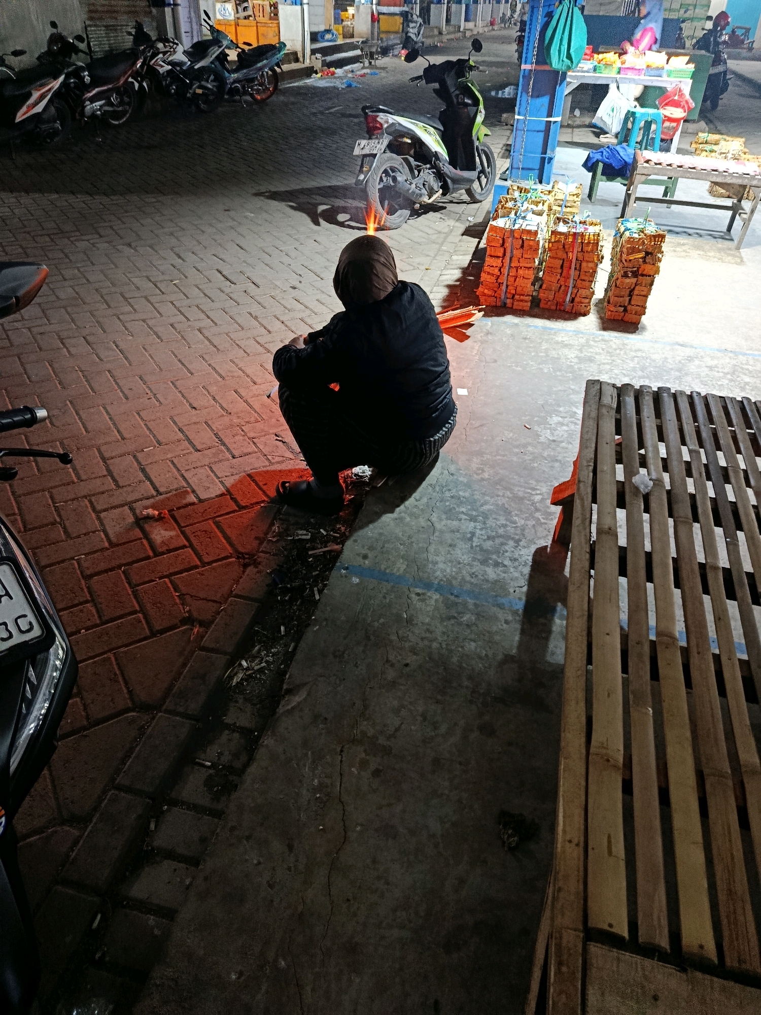 A person is crouching on a sidewalk at night, facing away from the camera. The person is wearing a dark hooded garment and dark pants. In front of the person, there is a small, glowing red object, possibly a firework or a burning cigarette, casting a warm light on the person's legs and the brick pavement. To the right of the person is a wooden bench made of bamboo slats. In the background, there are several parked motorcycles, some market stalls with goods on display, and a blue structure. The scene appears to be an outdoor market or street vending area. The license plate on the motorcycle partially visible on the left reads "M 905 MB". The pavement is made of rectangular red bricks. The overall lighting suggests it is nighttime.