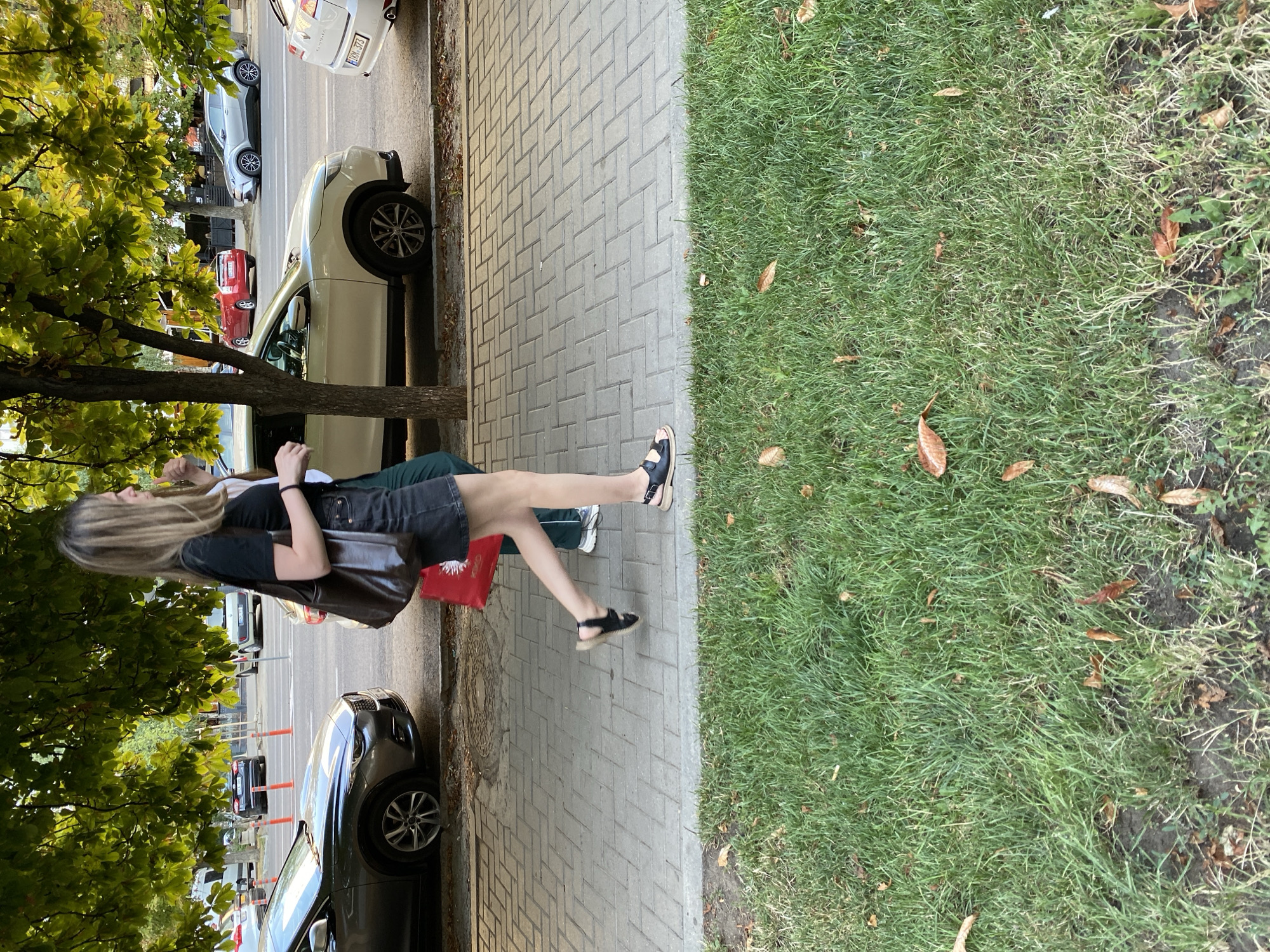 A young woman with blonde hair, wearing a black t-shirt, denim shorts, and black sandals, is walking on a sidewalk in Chișinău, Moldova. She is carrying a large brown shoulder bag and a red shopping bag. To her right, a person in a green shirt is partially visible. They are walking on a sidewalk made of light grey interlocking pavers. The sidewalk is adjacent to a street with several parked cars, including a dark grey sedan on the left, a white SUV in the middle, and a white hatchback on the right. Trees with green leaves overhang the sidewalk and street. In the foreground, a patch of green grass with scattered brown leaves is visible. The scene appears to be an urban street environment.