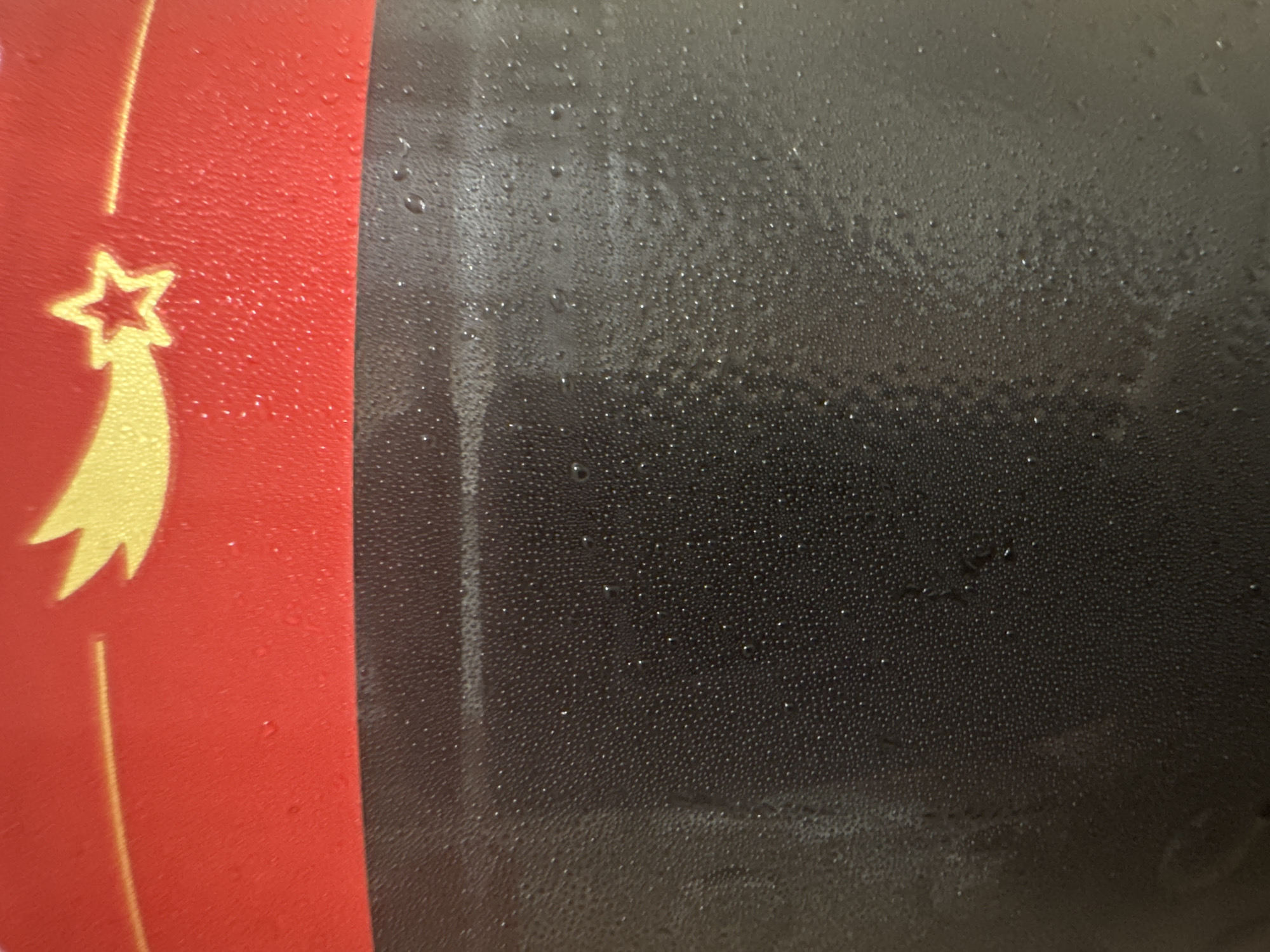 The image is a close-up, abstract shot of a dark, textured surface, possibly plastic or a coated metal, covered in condensation droplets of various sizes. The upper portion of the image features a red band with a yellow design resembling a shooting star or comet with a smaller star within it. A thin yellow line is also visible on the red band. The condensation is most prominent on the dark surface, creating a speckled, slightly blurred appearance with highlights from the droplets. The lighting suggests an indoor environment with a light source reflecting off the condensation. No specific location in the city of Unknown, Unknown can be determined from the image.