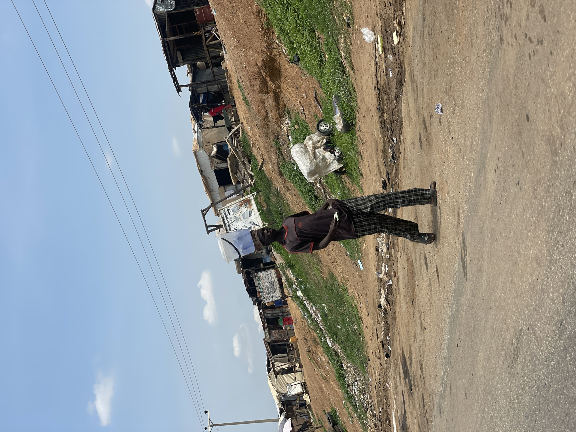 The image depicts a street scene in Kwali, Nigeria. An individual stands on the right edge of a paved road, wearing a dark shirt and patterned pants. Along the left side of the road is an area characterized by a mix of vegetation and trash, adjacent to a series of makeshift dwellings or shacks constructed from various materials. Power lines are visible against a blue sky with scattered clouds.