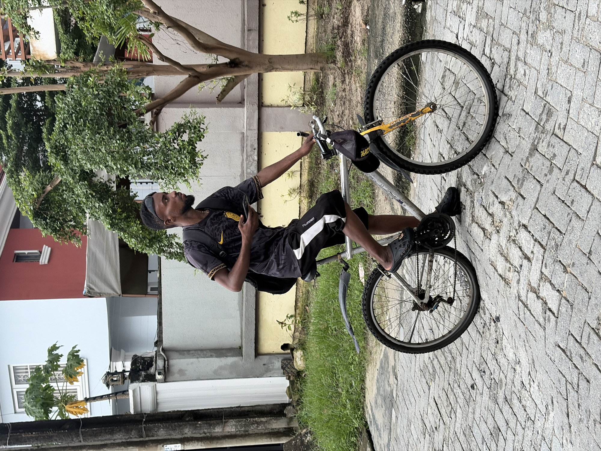 A full shot shows a young Black man with a beard and a short haircut, wearing sunglasses on his head, riding a bicycle down a brick-paved street in Igboefon, Nigeria. He is wearing a black t-shirt with a yellow and black abstract design, black shorts with a white stripe, and grey slides. He has a black backpack on his shoulders and is holding a yellow cell phone in his right hand. The bicycle is silver with black tires and a black fender over the rear wheel. A black bag hangs from the handlebars. Behind the man is a light blue and red building with white trim and windows. There are also fences, trees, and grass visible in the background. The overall lighting suggests an overcast day.