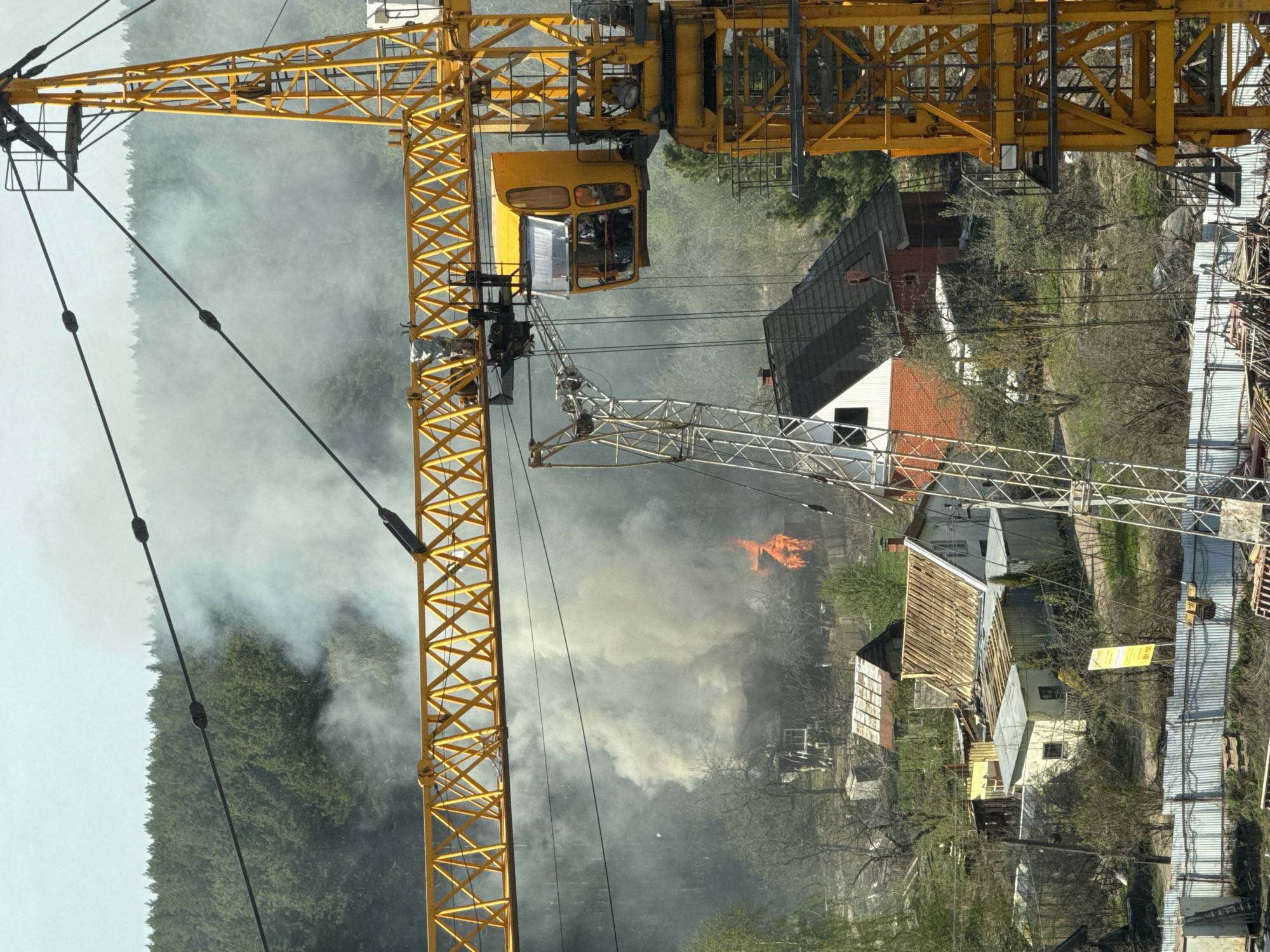 The image shows a fire in a residential area of Izhevsk, Russia. Thick smoke obscures much of the background, but flames are visible amidst the smoke. A yellow tower crane stands prominently in the foreground on the left side of the image. The view appears to be from an elevated position, looking down on the scene. In the background, there are trees and several houses.