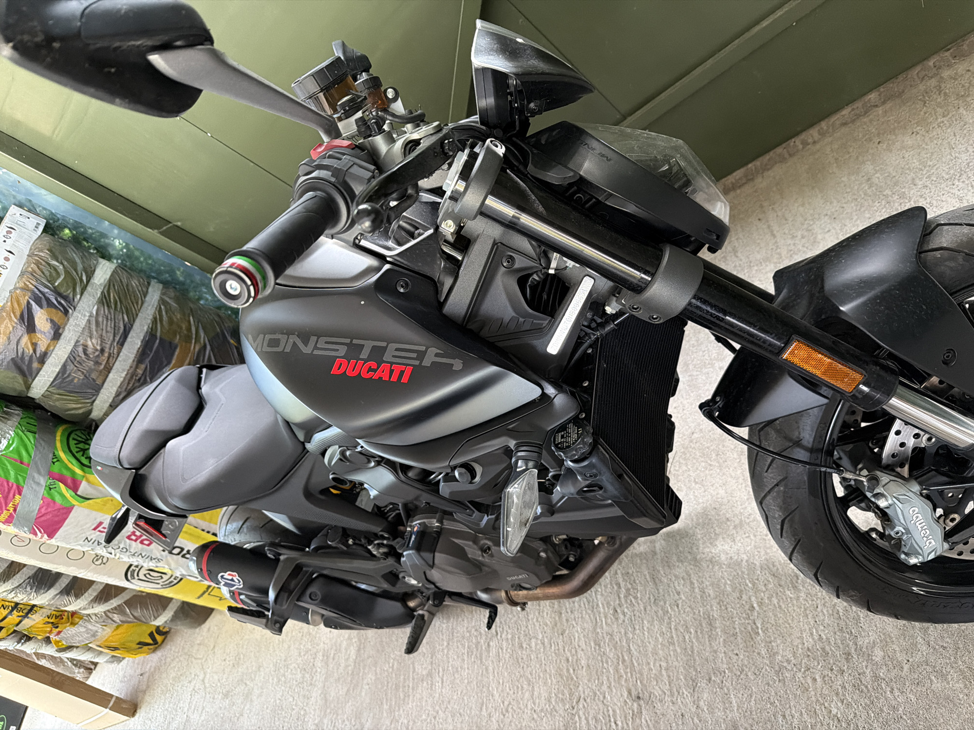 The image shows a black Ducati Monster motorcycle parked indoors, likely in a storage area or garage, in Sofia, Bulgaria. The motorcycle is the primary subject, viewed from a slightly elevated angle, facing towards the right of the frame. It is dark in color with red "DUCATI" lettering on the fuel tank and a black exhaust pipe with a red and black tip. Visible components include the front wheel with a silver Brembo brake caliper, the suspension forks, handlebars with grip ends featuring green, white, and red stripes, the seat, and the rear tire. In the background, there are stacks of rolled materials, possibly insulation or carpet, wrapped in plastic and paper. The floor appears to be concrete. The overall lighting suggests an indoor setting with some ambient light from an unseen source.