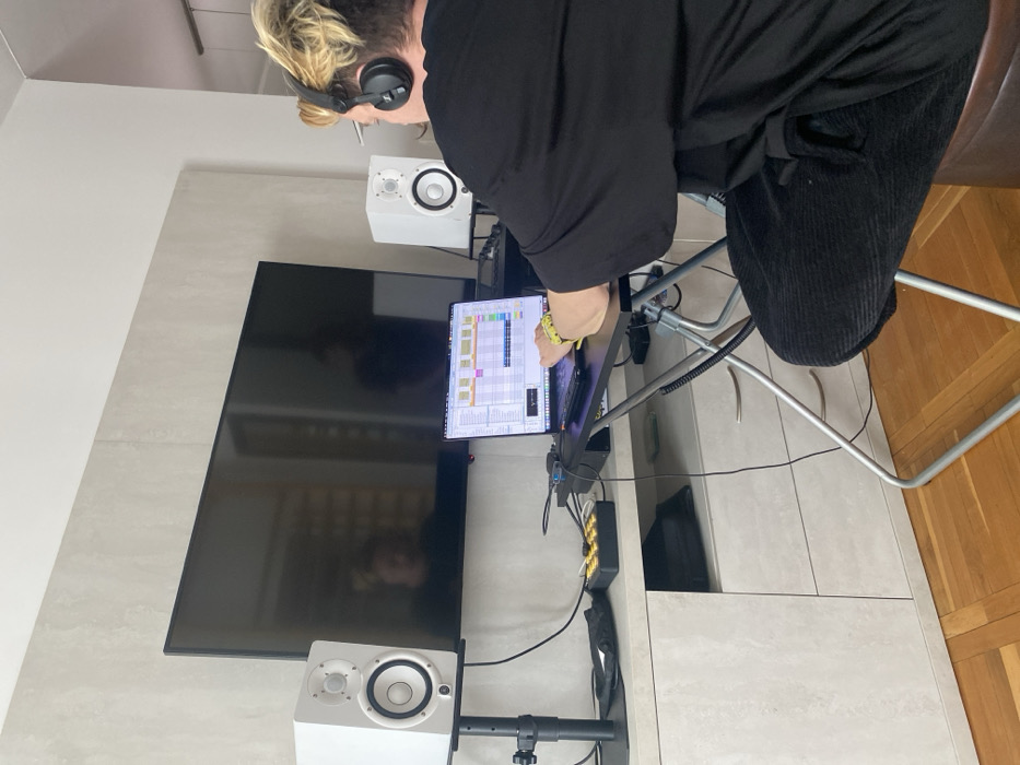 The image displays an indoor scene located in the city of Vozdovac Urban Municipality, Serbia. An individual, viewed from behind, sits at a black folding desk, engaged in music production. The person wears a black oversized t-shirt, black corduroy-like pants, and black over-ear headphones. A yellow wristband is present on their left wrist.
The central focal point is a laptop computer displaying music production software (identified as Ableton Live by its interface), which the individual is actively operating with their right hand on the keyboard/trackpad. The laptop is positioned on the black folding desk, which has silver metal legs. Directly behind the laptop, a large black flat-screen television is mounted on a light grey wall. Reflections of the room and a partial figure are visible on the television screen.
Two white studio monitor speakers (Yamaha HS series) stand on black adjustable stands, flanking the television. A black audio interface or USB hub is connected to the laptop, with cables visible behind the equipment. To the left of the laptop, on a light grey media console, sits a small black MIDI controller with 16 illuminated yellow pads. The media console features two drawers and an open compartment on the left, which contains a black bag. The floor is light brown parquet.